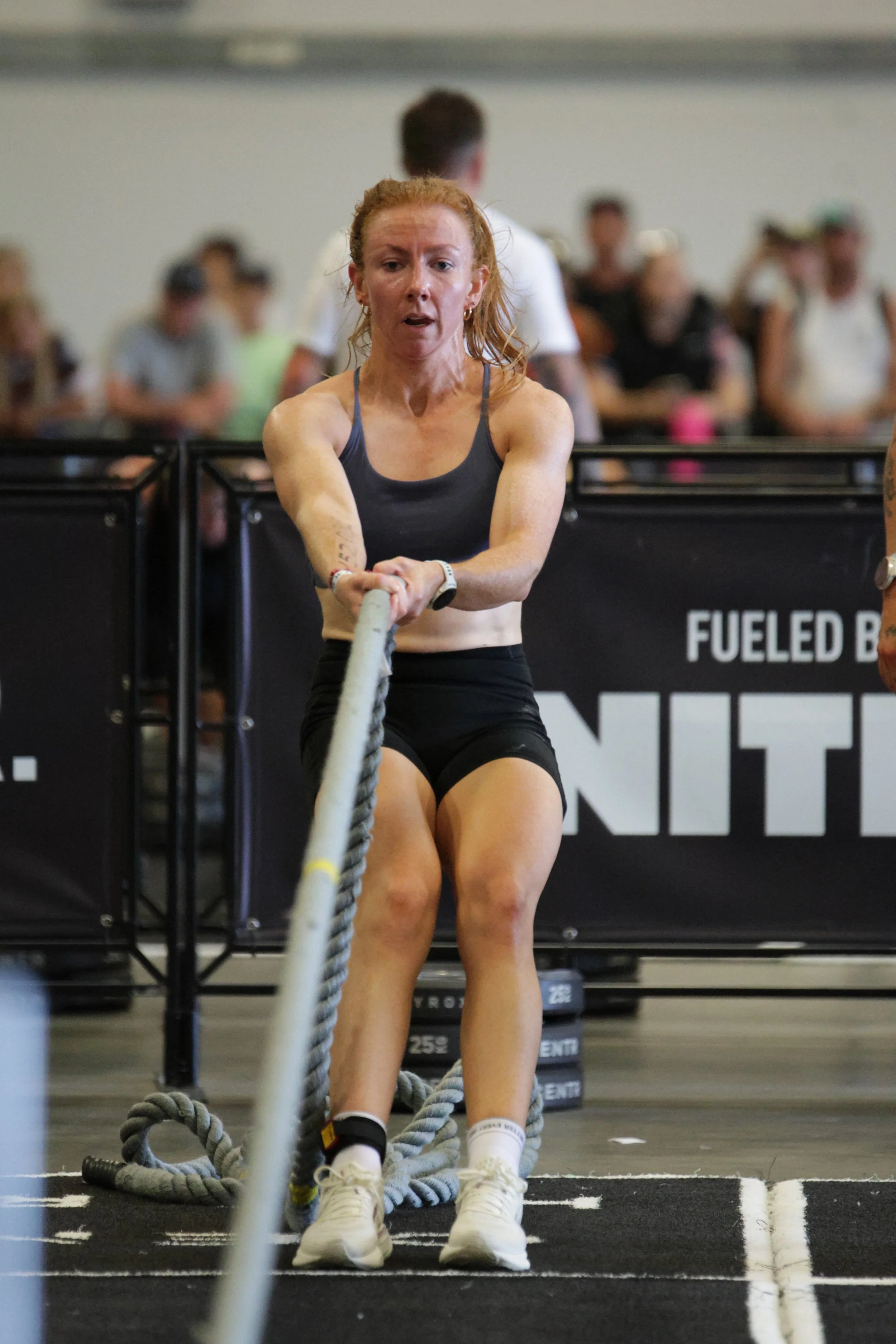 A woman with red hair in athletic attire pulling a heavy rope during a fitness competition in a gym, with a blurred audience in the background.
