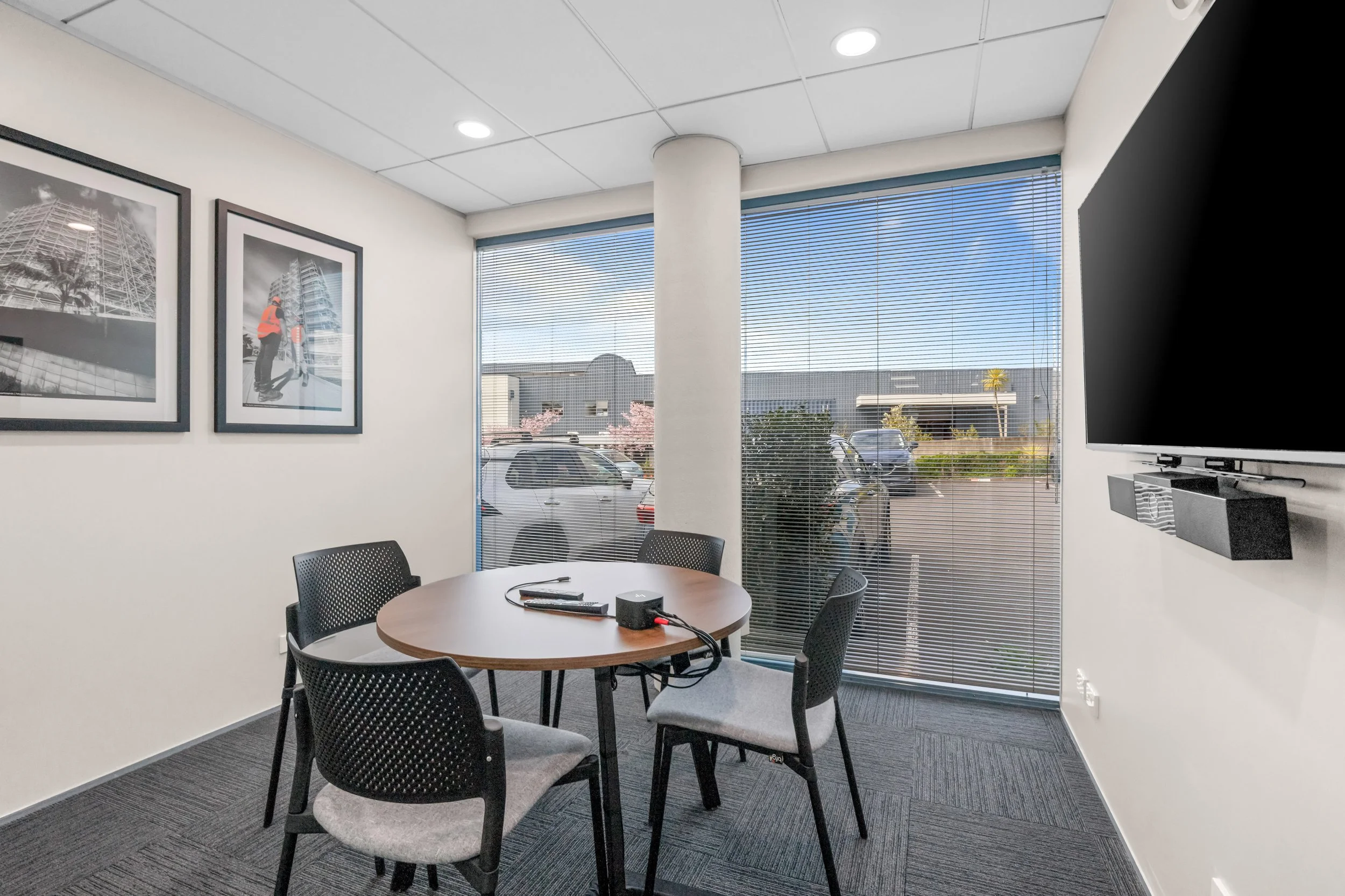 A conference room with a round wooden table surrounded by five black chairs with grey cushions, positioned on a dark carpet. On the table are some electronic devices and cables. The room has white walls with two framed black-and-white photographs of 