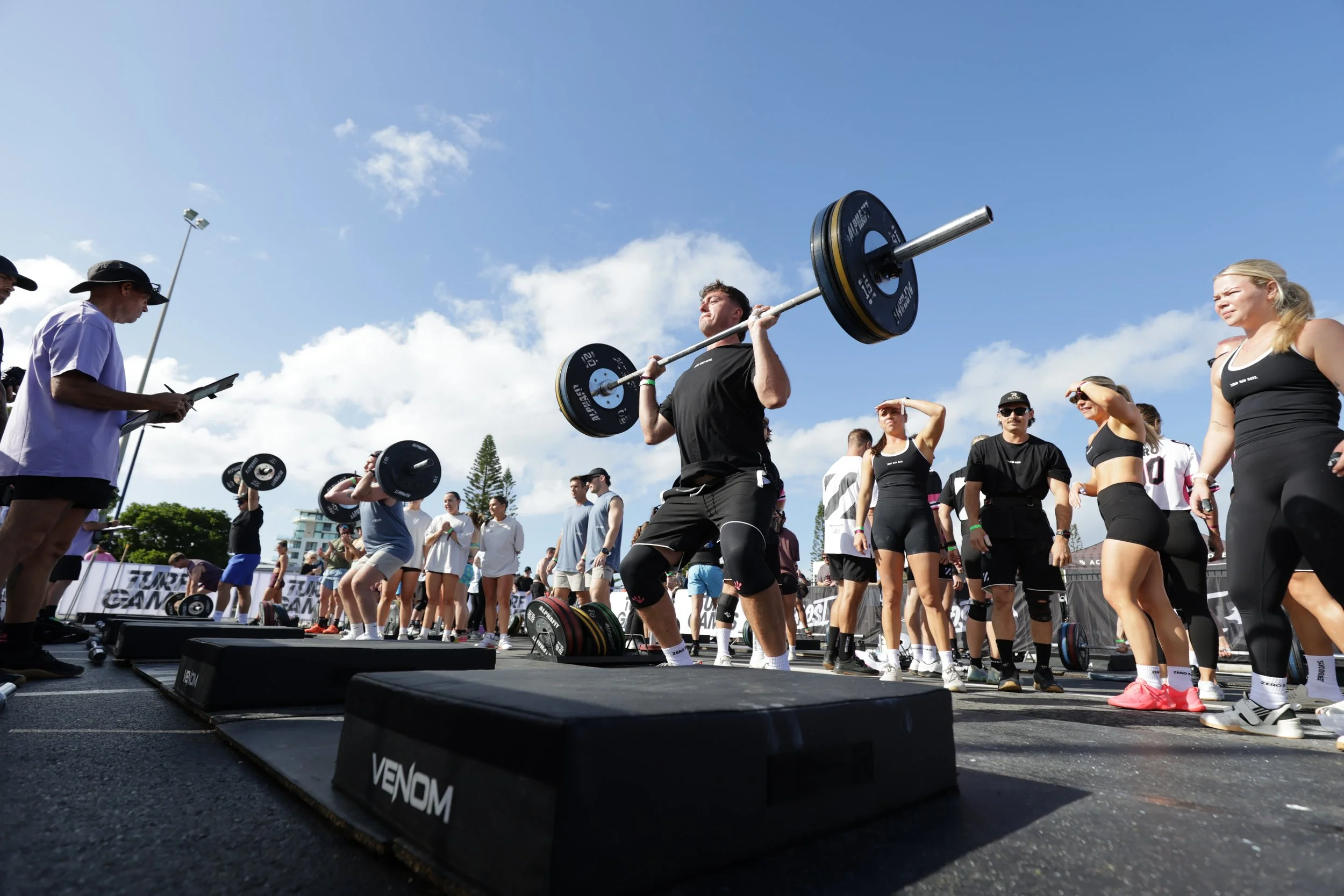 A man lifting a barbell during a fitness competition or outdoor workout event with a group of people watching, some holding phones or clipboards, in an open area with a bright blue sky and scattered clouds.