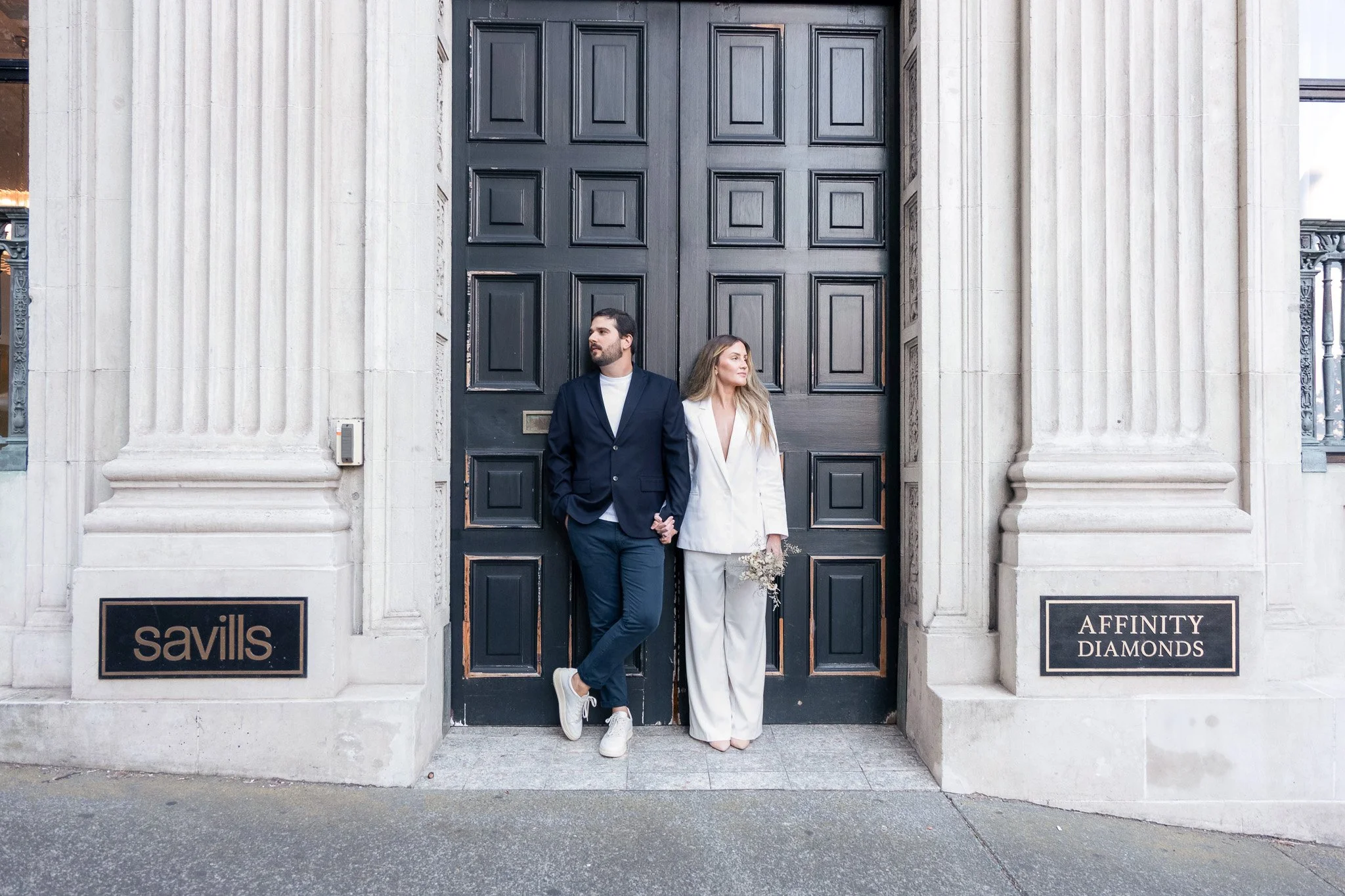 A man and woman standing in front of a large black door with white stone columns on either side. The man is wearing a navy suit with white sneakers, and the woman is dressed in a white outfit holding a bouquet of flowers. There are black signs with g