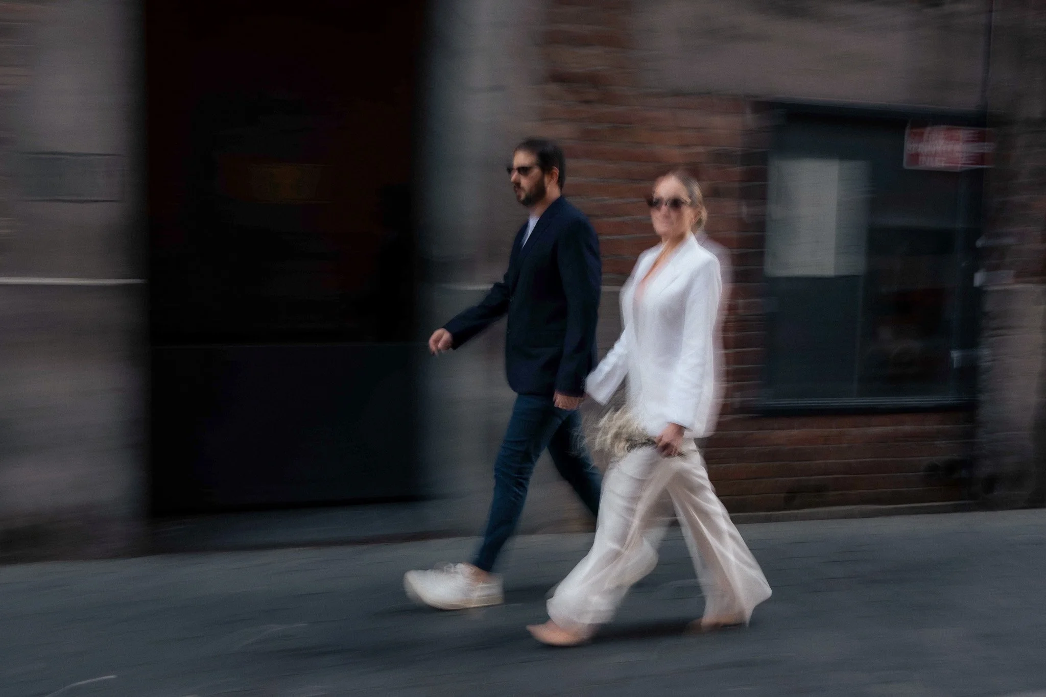 A man and a woman walking together on a city sidewalk, both wearing sunglasses and stylish clothing. The man is in a dark blazer and jeans, and the woman is in a white outfit with feathered details on the pants. They are walking past a brick building