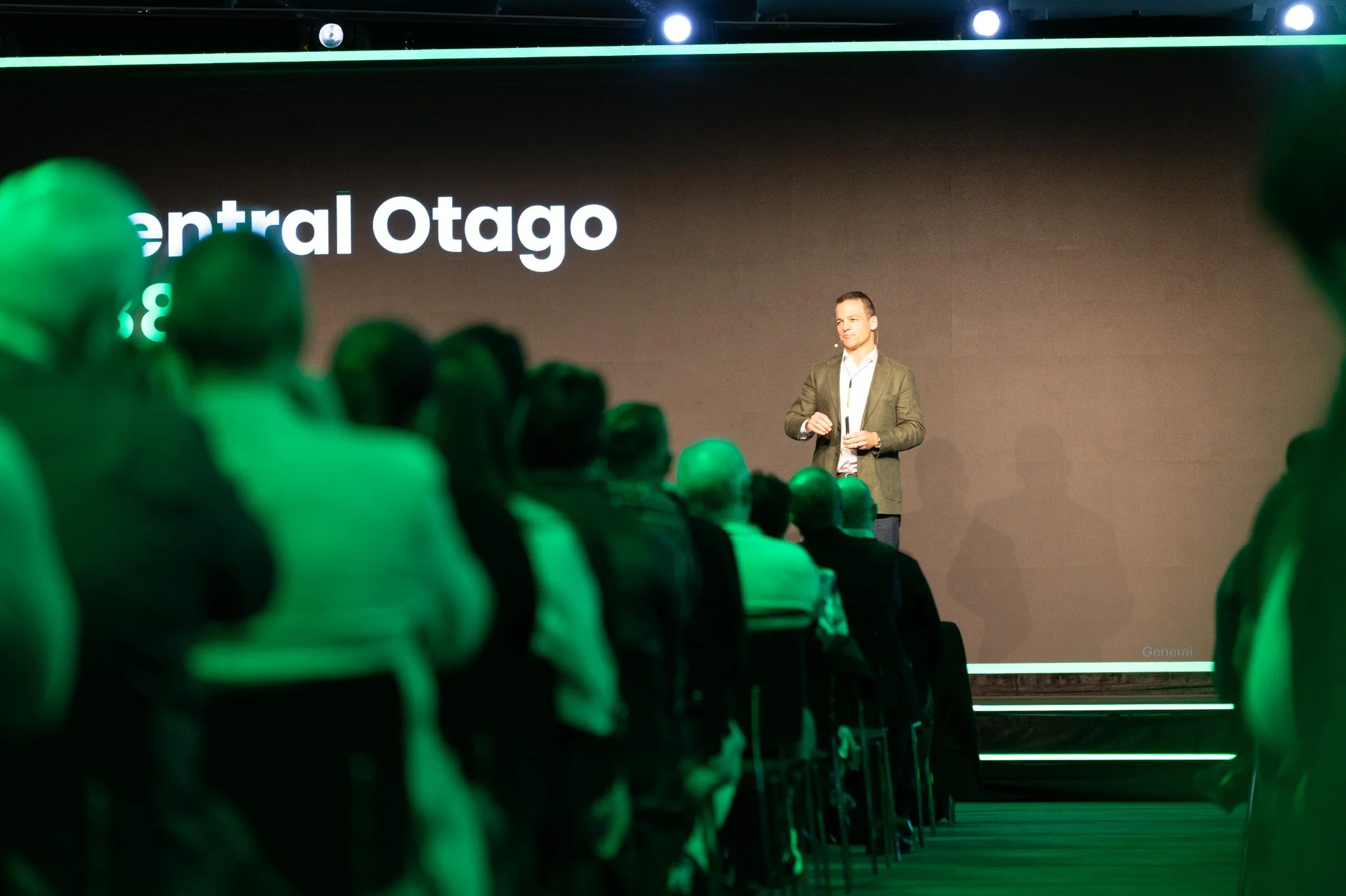A man in a blazer giving a presentation on stage, with a large screen behind him displaying text and a row of green lights above. Audience members are visible in the foreground.