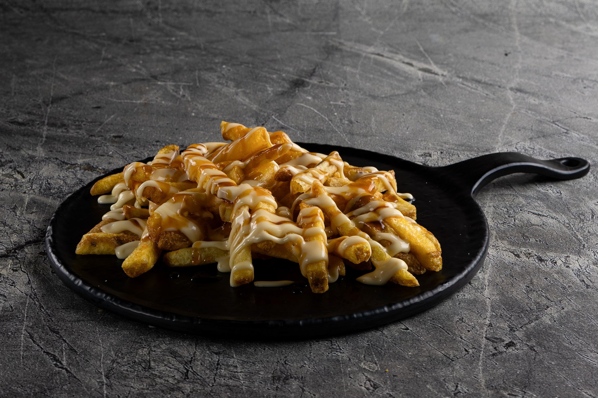 A black plate of cheese fries topped with melted cheese, drizzled with white and caramel-colored sauces, on a textured dark gray surface.
