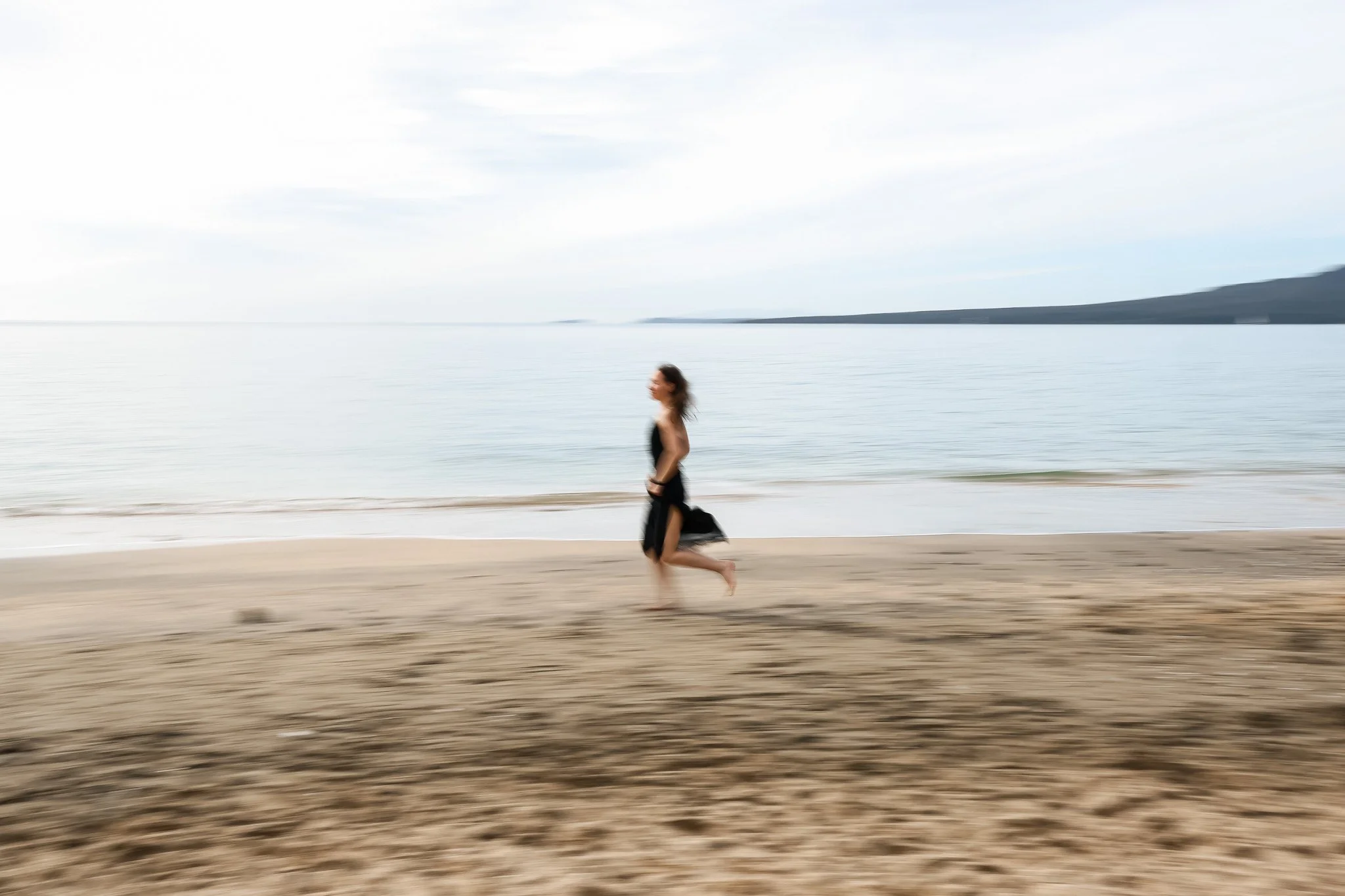 A person running on the beach near the water, with a distant landmass and cloudy sky in the background.