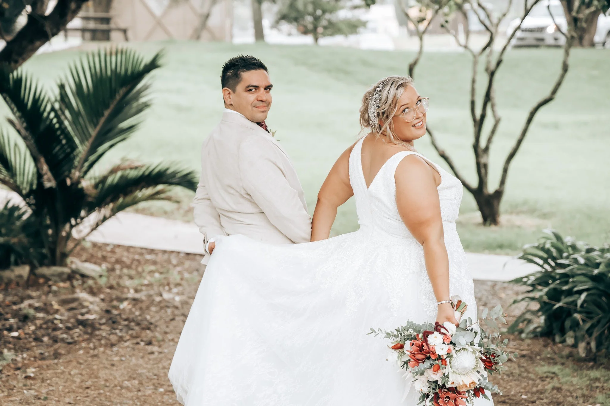 Bride and groom at a wedding outdoors, with the bride holding a bouquet of flowers and the groom standing behind her, both smiling and looking at the camera. The bride is wearing glasses and a white wedding dress, while the groom is dressed in a ligh