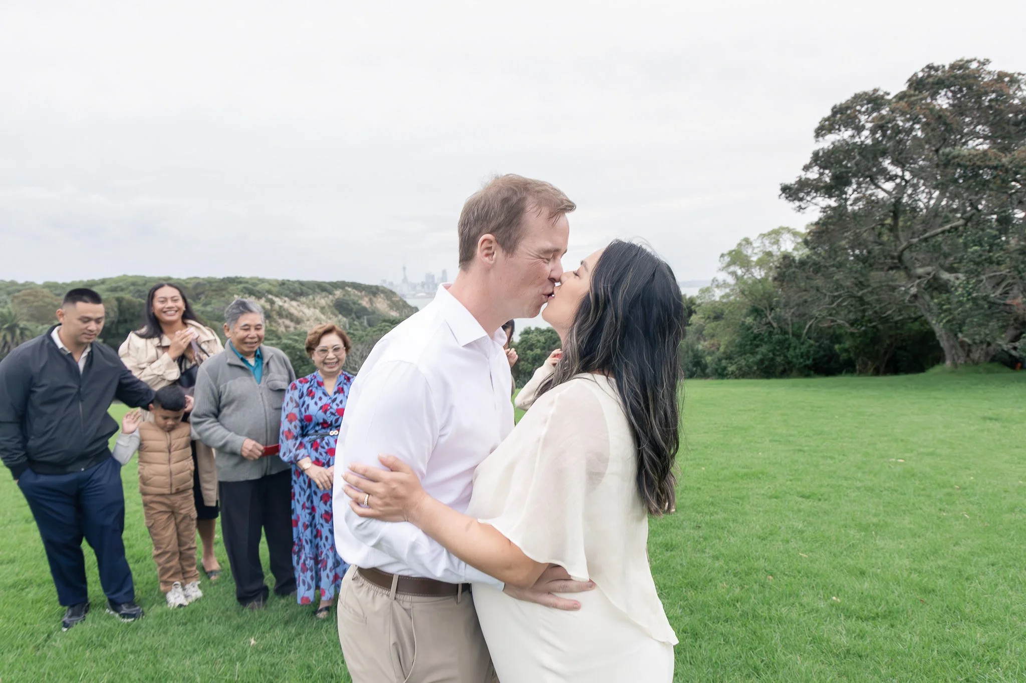 A man and woman sharing a kiss outdoors, with a group of people in the background on a green field and trees, overcast sky.