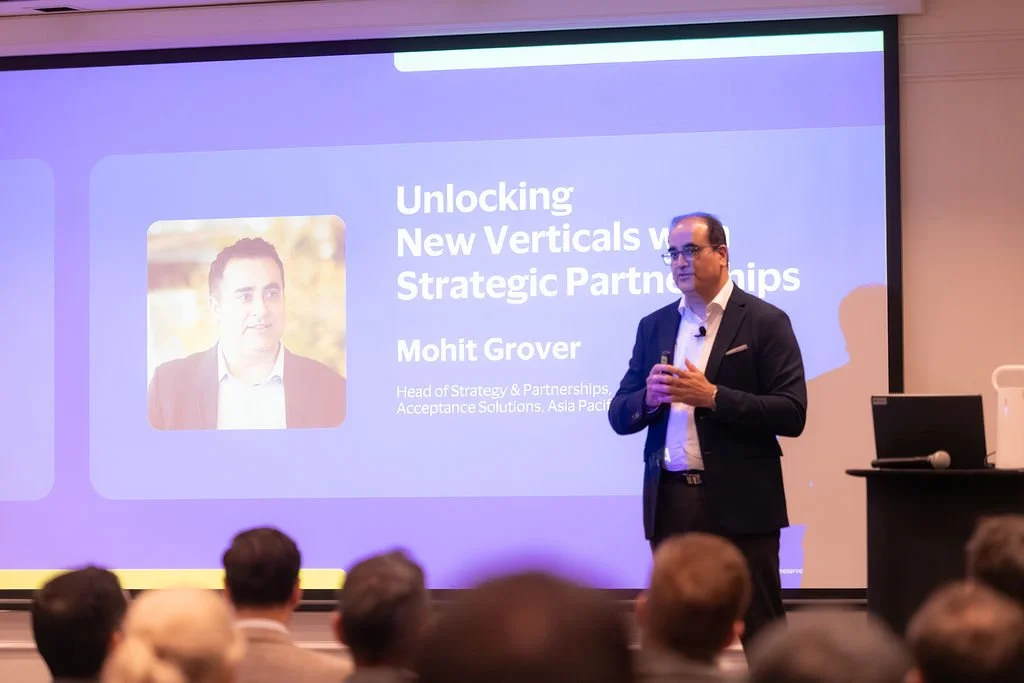 A man in a suit presenting at a conference, with a slide on the screen behind him titled "Unlocking New Verticals w/ Strategic Partnerships" by Mohit Grover, Head of Strategy & Partnerships at Acceptance Solutions, Asia Pacific.