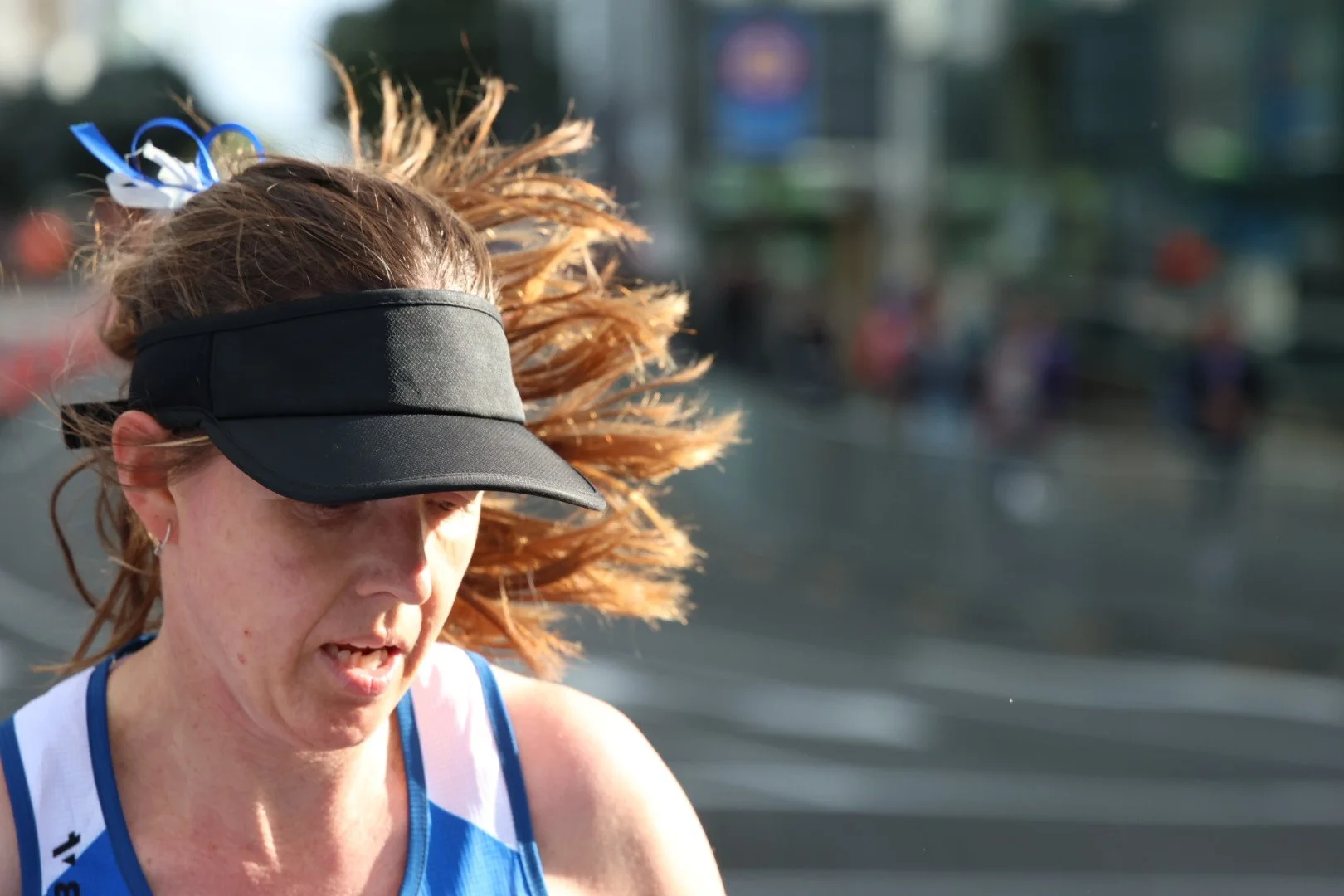 Close-up of a woman with red hair, wearing a black visor and a blue and white sports top, running outdoors on a sunny day.