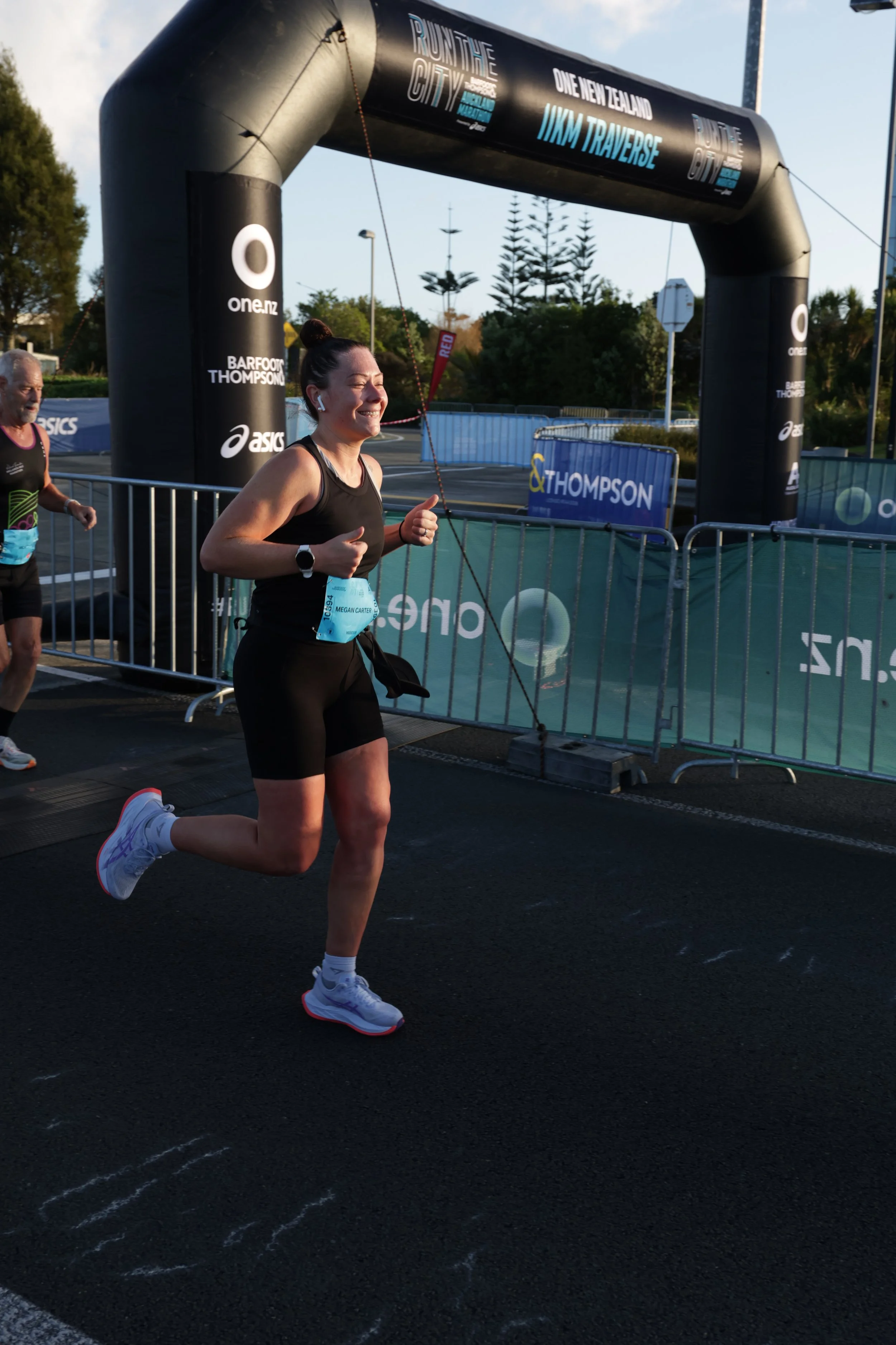 A woman running in a race, crossing the finish line with a smile, wearing black athletic gear, white running shoes, and wireless earbuds during a sunny day.