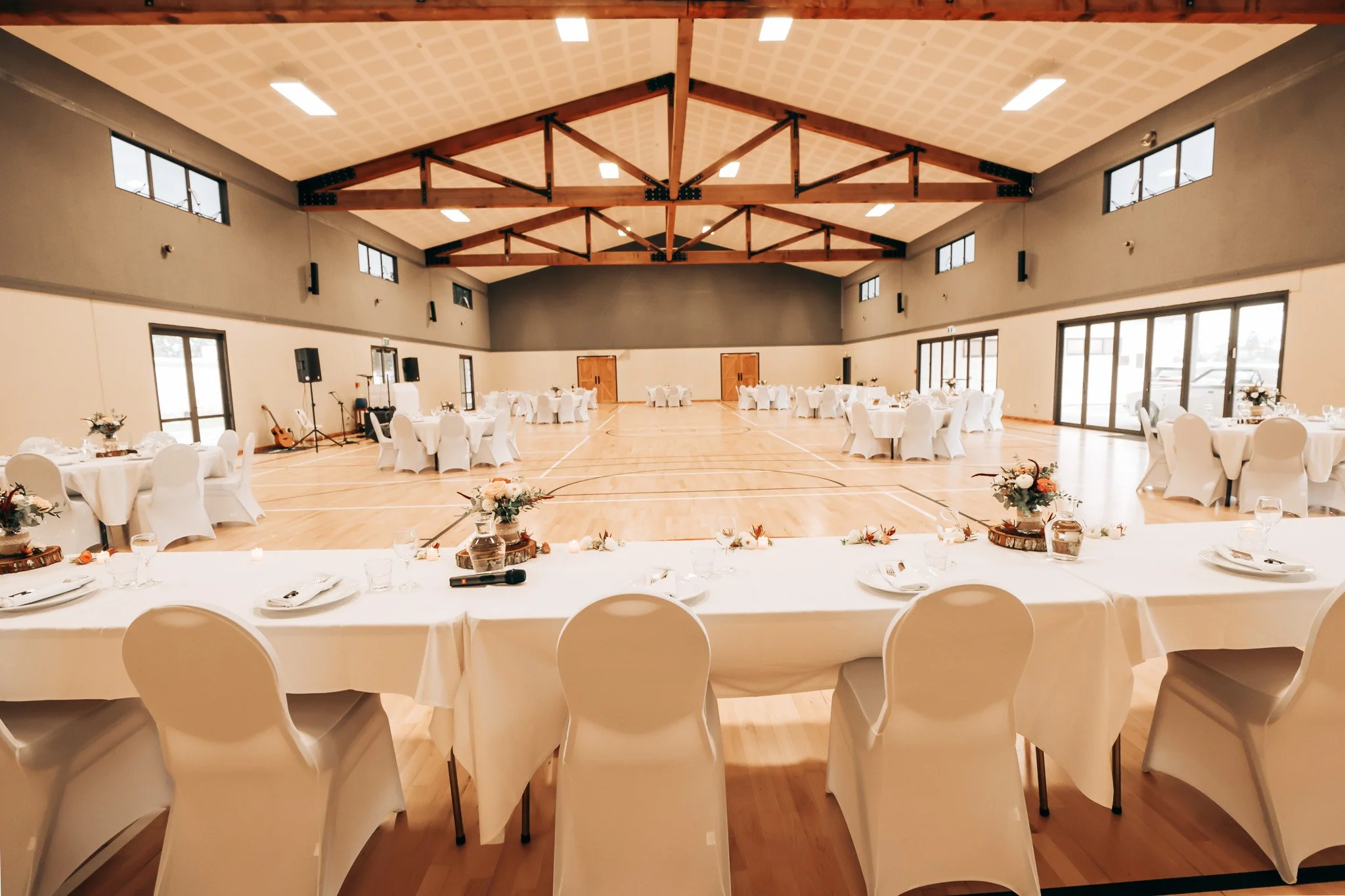 A banquet hall set up for a wedding reception with round tables covered in white tablecloths, decorated with floral centerpieces, and white chairs. The hall has a high ceiling with wooden beams, windows along the upper walls, and a wooden floor.