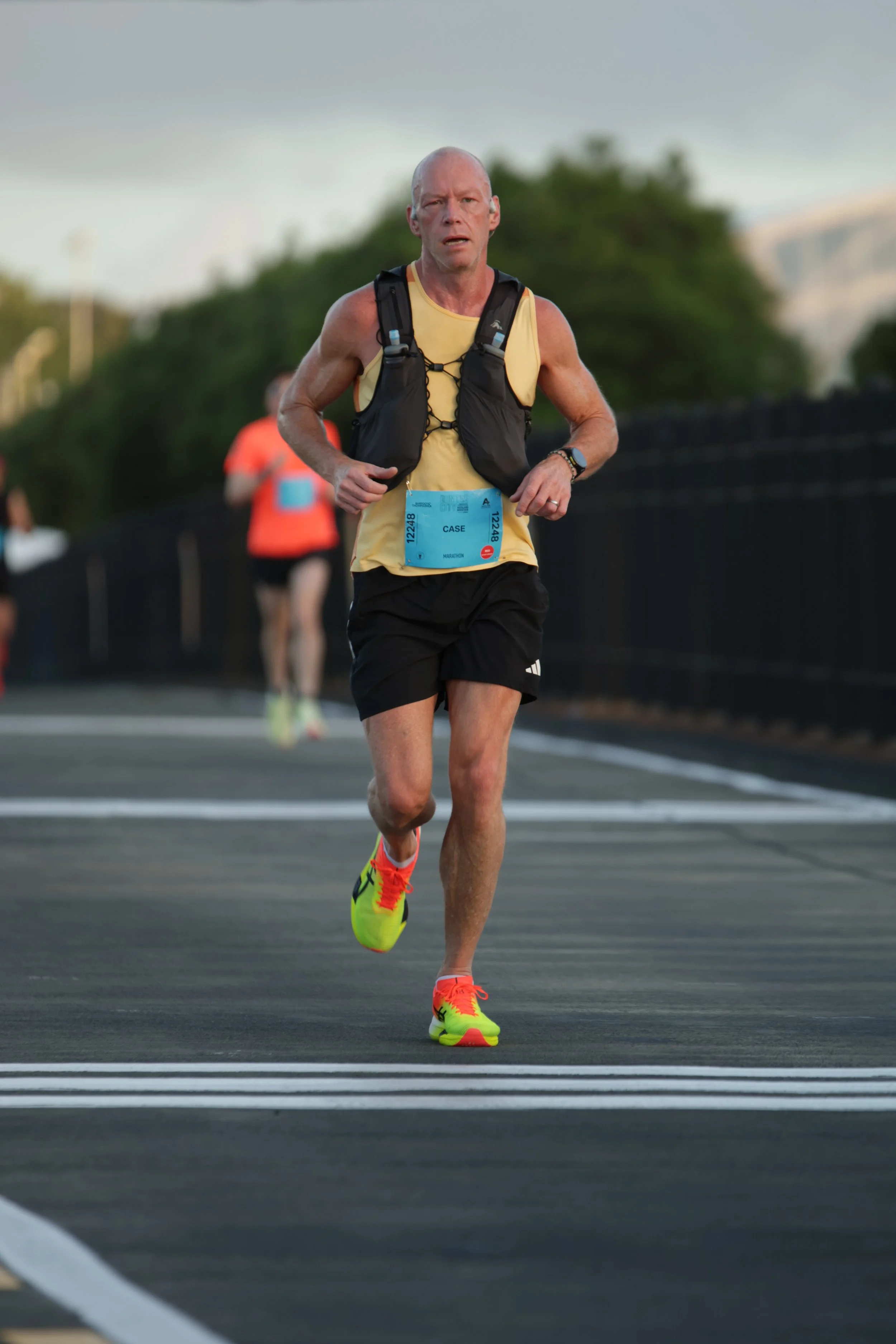 A male marathon runner wearing a yellow sleeveless shirt, black shorts, and bright yellow running shoes, running on a paved road during a race.