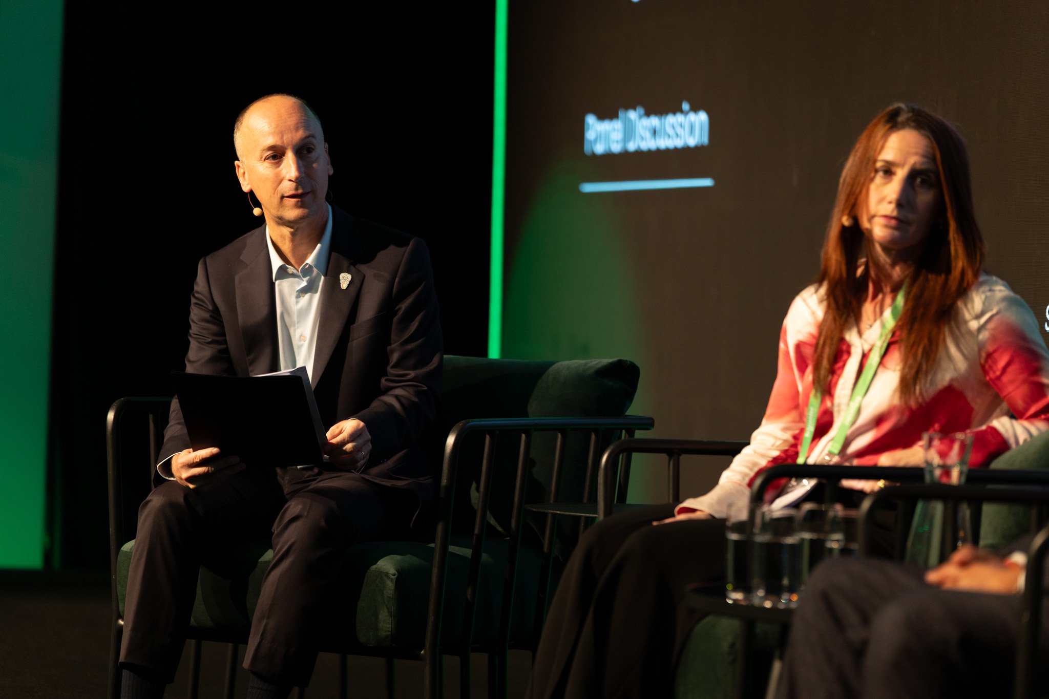 A man in a suit and a woman with long red hair sitting on stage during a panel discussion. The man has a microphone and is holding some papers, while the woman is looking forward. There is a large digital screen behind them displaying the words "Pane
