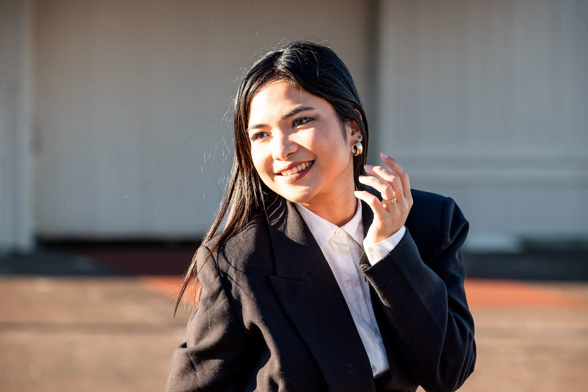 A young woman with dark hair smiling and wearing a black blazer and white shirt, standing outdoors in morning or late afternoon sunlight.
