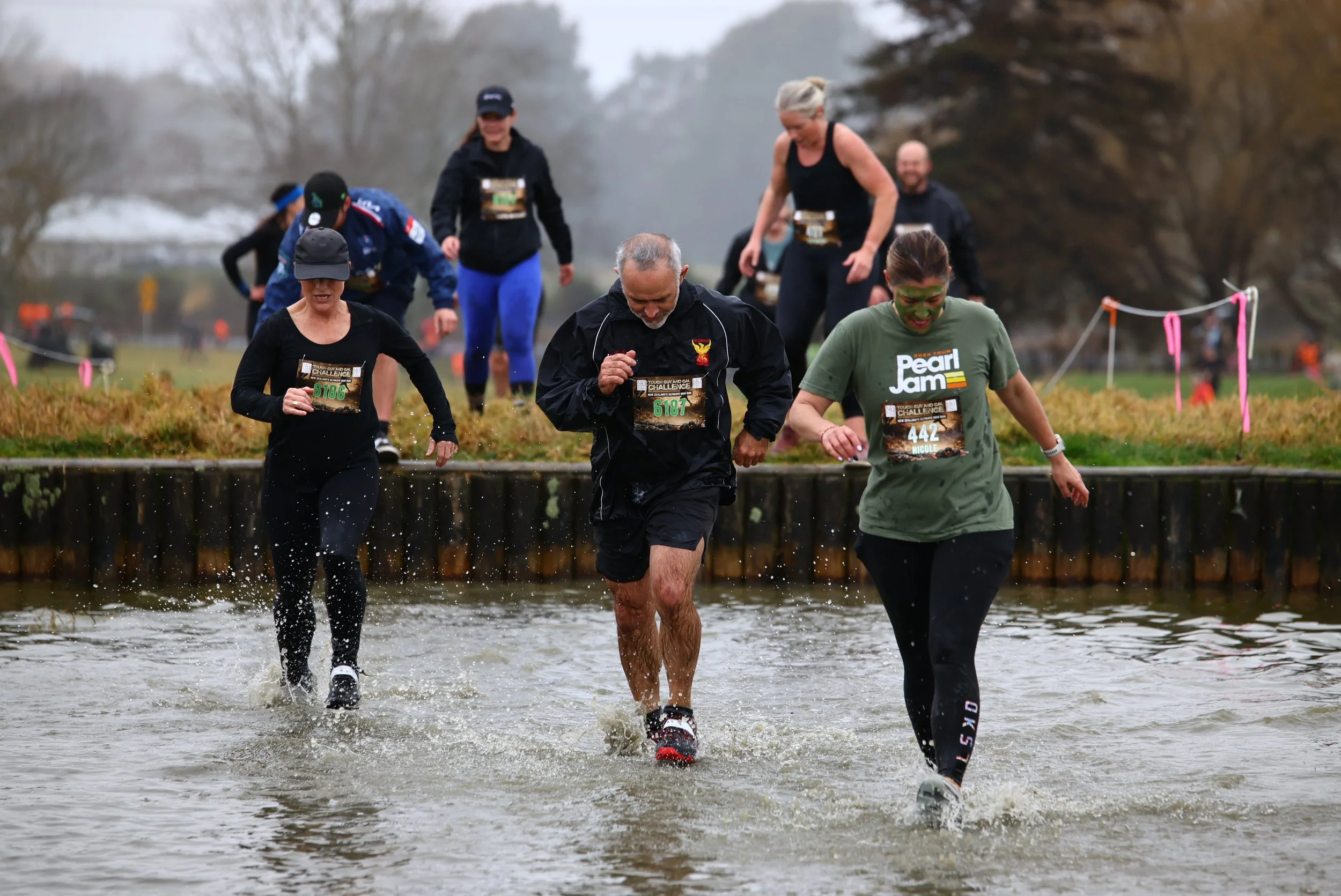 People participating in a trail run, crossing a shallow water obstacle during an outdoor race on a cloudy day.