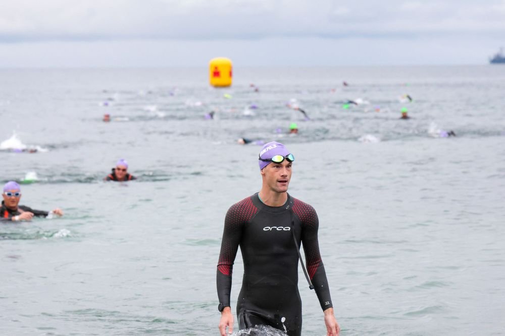 Swimmers participating in an open water race, with a male swimmer in the foreground wearing a black wetsuit and purple swim cap, and other swimmers swimming in the background near a yellow buoy.