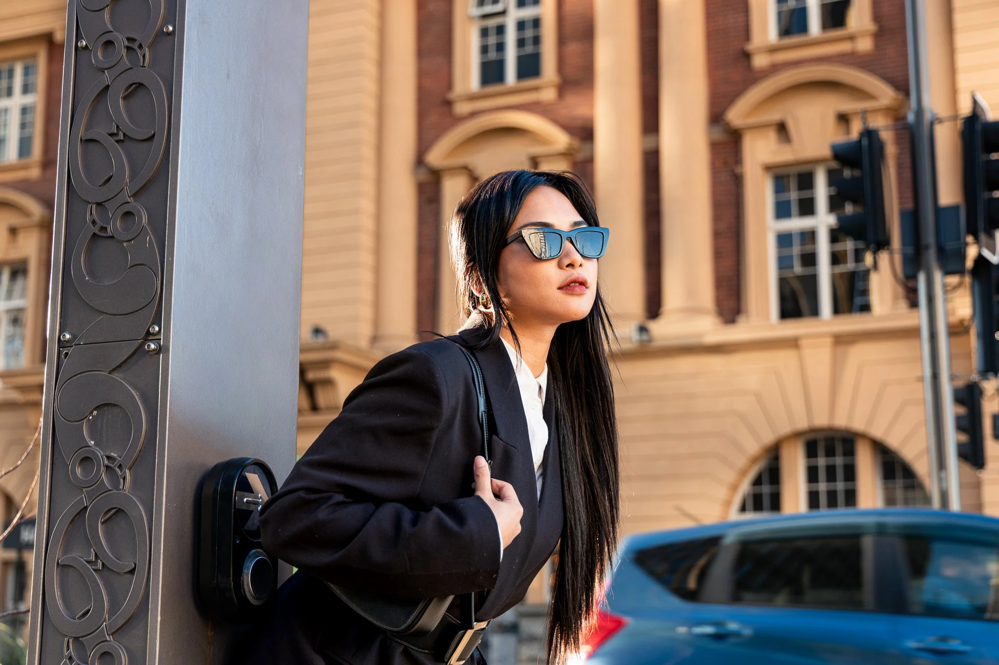 A woman with long dark hair wearing sunglasses, a black blazer, and a white shirt, standing on a city street near traffic lights and a building with detailed architecture.