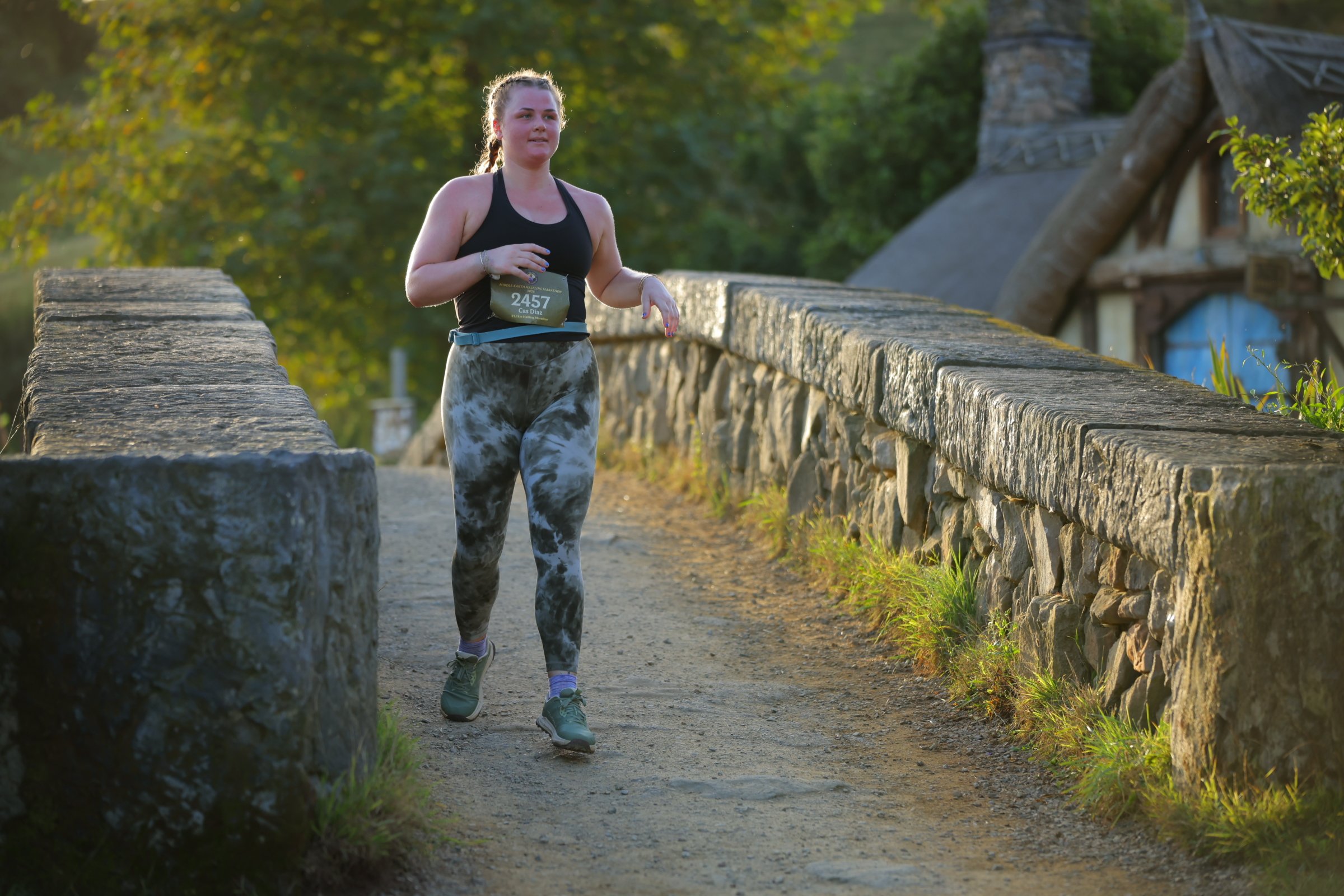 A young woman running outdoors on a trail next to a stone wall with trees and a house in the background, wearing a black tank top and patterned leggings.