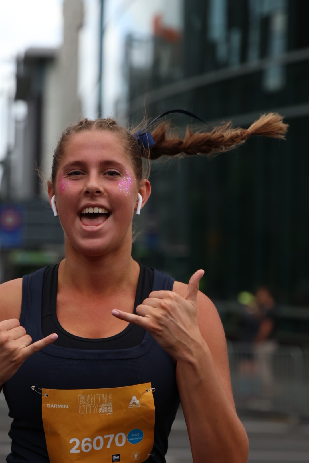 Woman smiling and wearing a race bib, making a fun hand gesture, outdoors in an urban area.