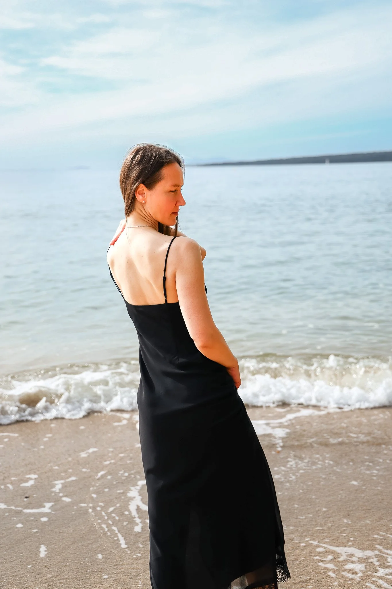 A woman in a black dress standing on the beach near the water with her eyes closed and looking downward.