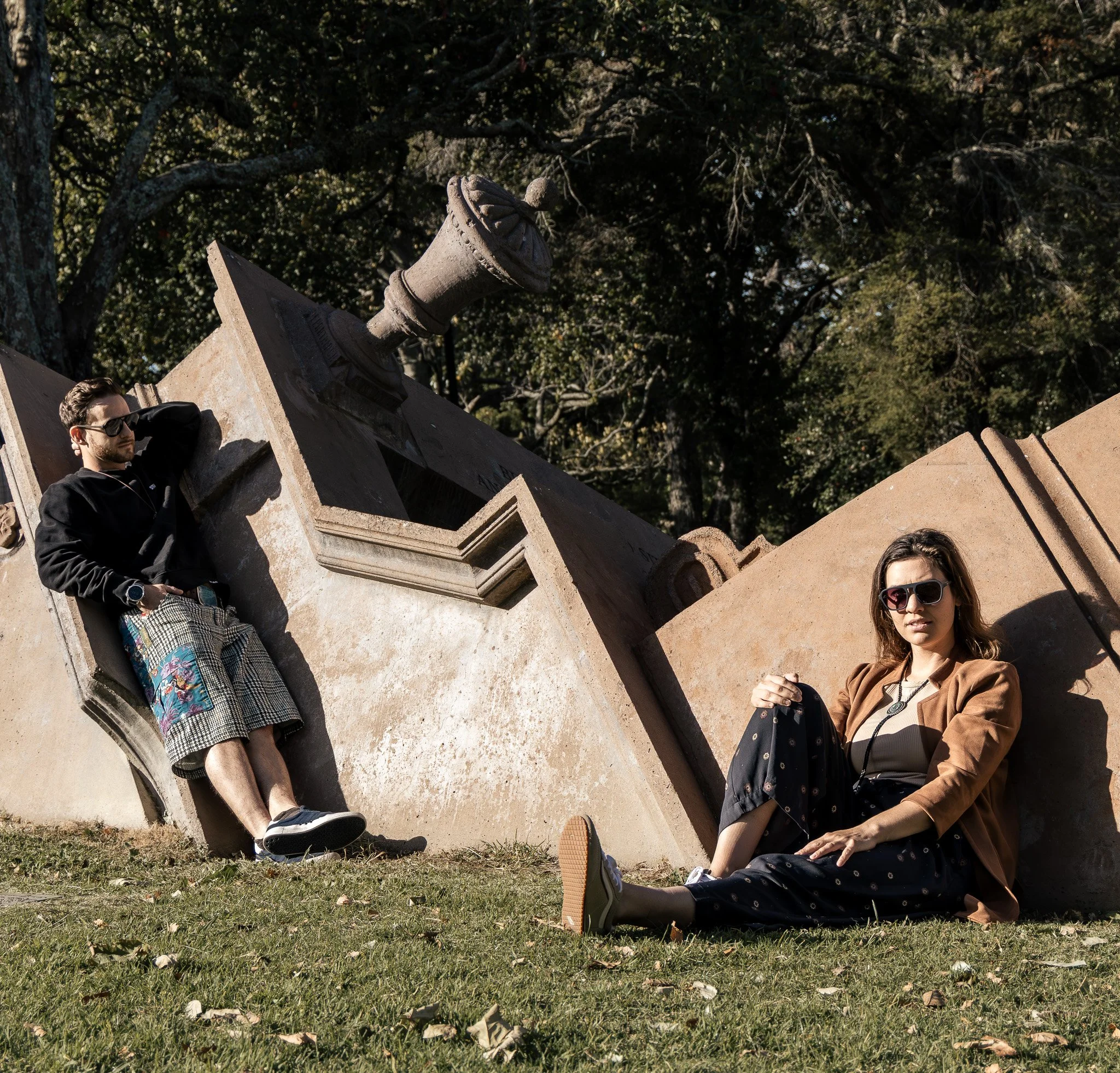 Young man wearing sunglasses, black hoodie, and checkered shorts sitting against a monument with an old cannon on top, while two young women sit nearby on the grass, one wearing sunglasses, a tan jacket, and black polka-dot pants, and the other stand