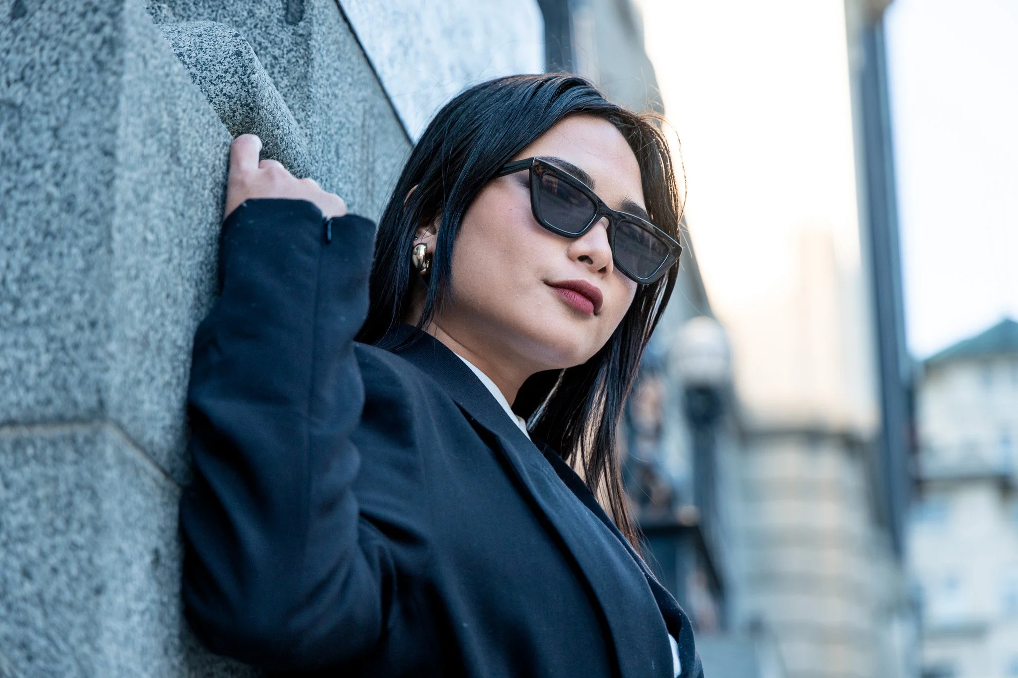 A woman with dark hair wearing sunglasses and a dark blazer, resting against a textured stone wall outdoors.