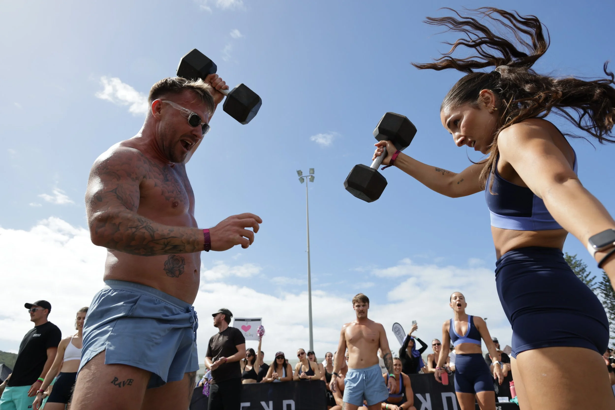Two people are team lifting dumbbells during an outdoor fitness event, with spectators and a clear sky in the background.