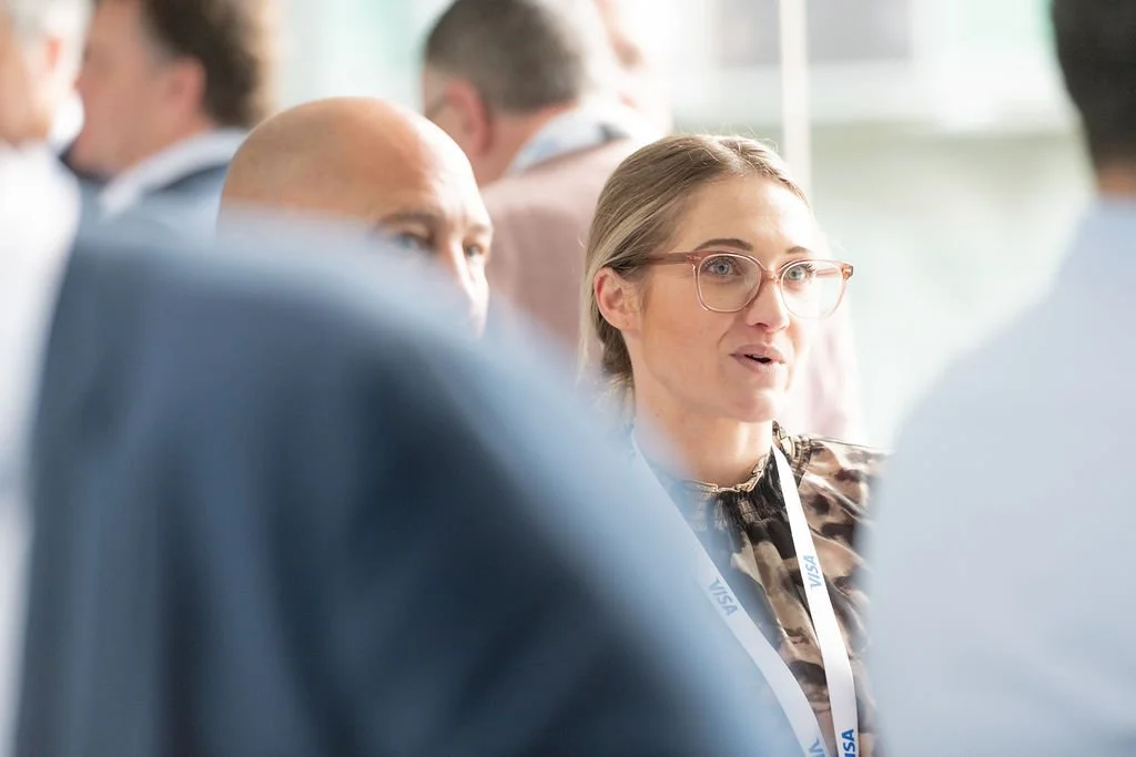 A woman with glasses engaged in conversation at a professional event, wearing a lanyard with a visa badge, surrounded by other attendees.