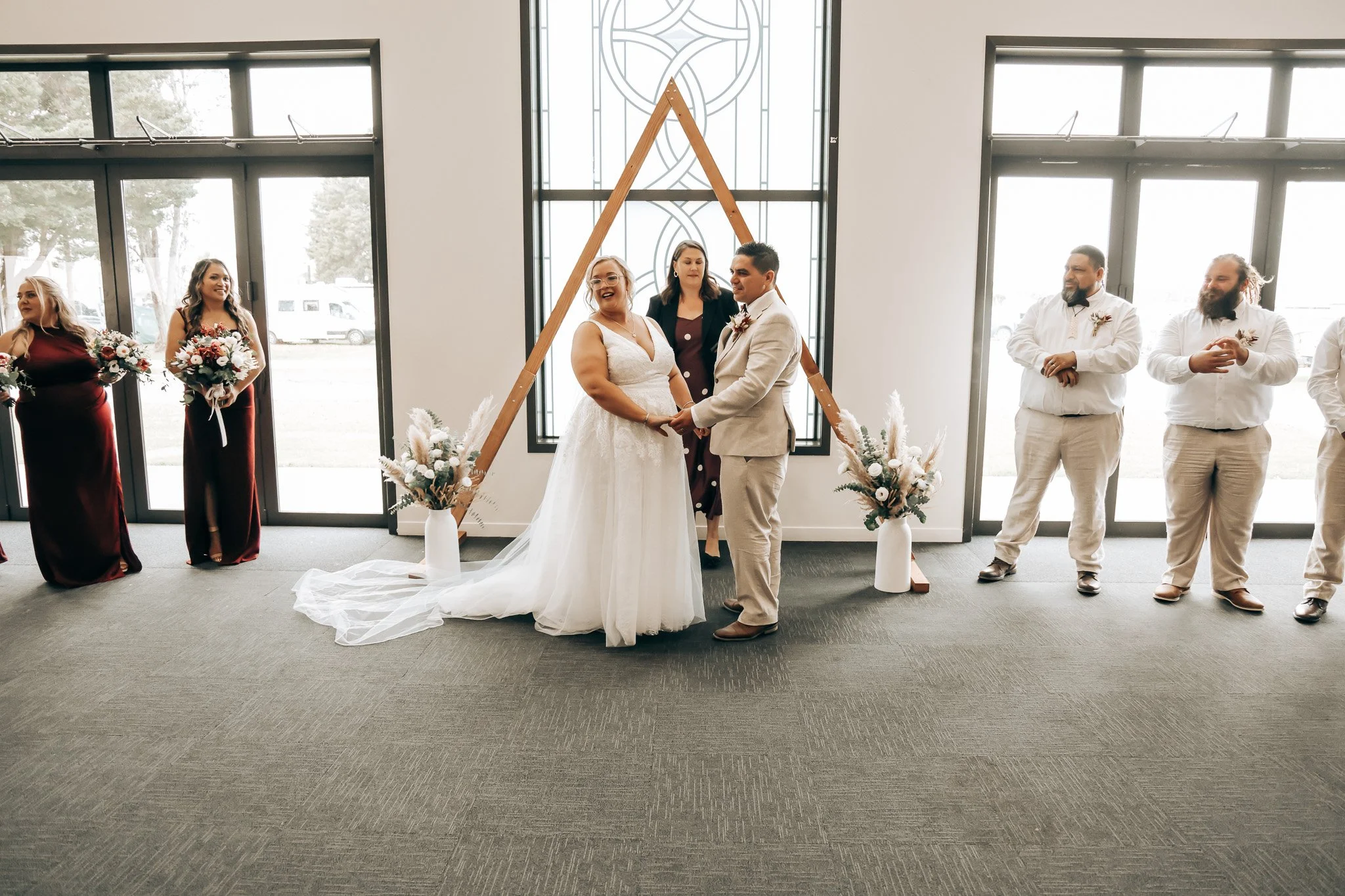 A wedding ceremony with the bride and groom holding hands and standing in front of a wooden triangular arch, surrounded by bridesmaids and groomsmen in a bright indoor space with large windows.
