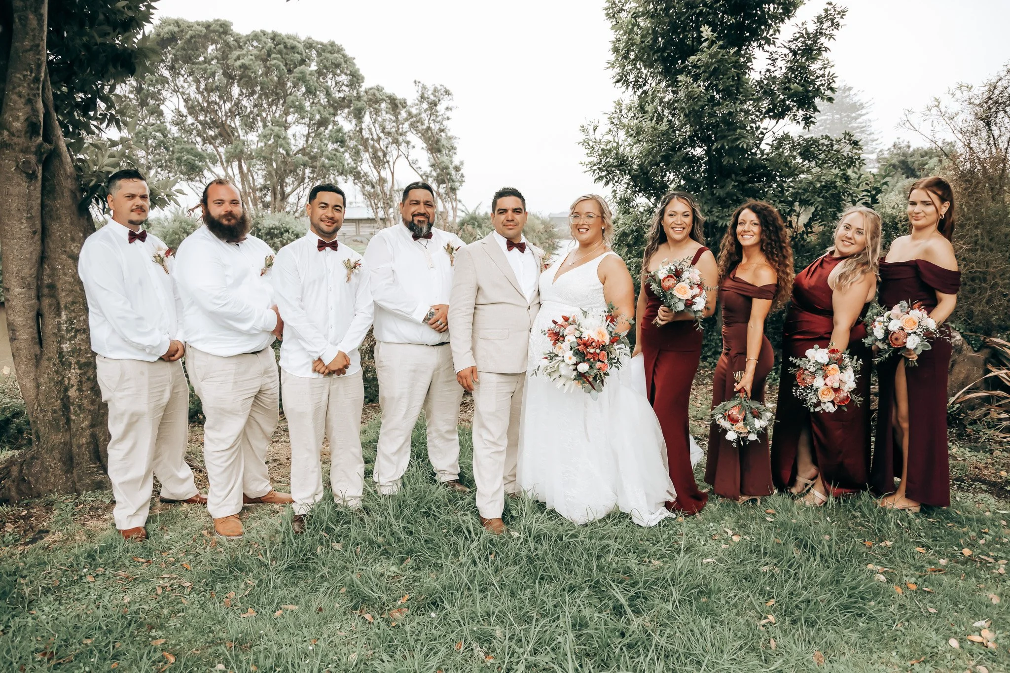 A group of ten people, including a bride and groom, standing outdoors on green grass with trees in the background, dressed in wedding attire.