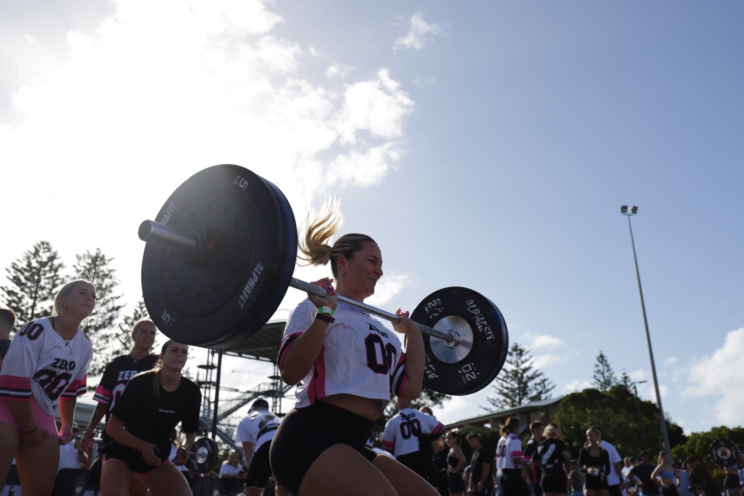 A woman lifting a barbell during a daytime outdoor fitness event, with other women and event structures visible in the background.