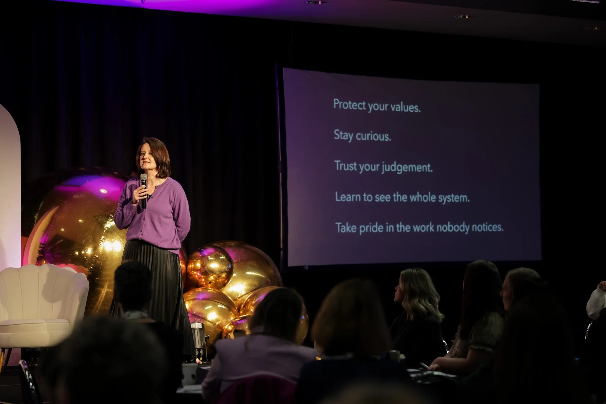 A woman in a purple sweater and black skirt speaking into a microphone at a presentation event. Behind her, a large screen displays motivational points, including 'Protect your values,' 'Stay curious,' and 'Trust your judgment.' The stage decorated w
