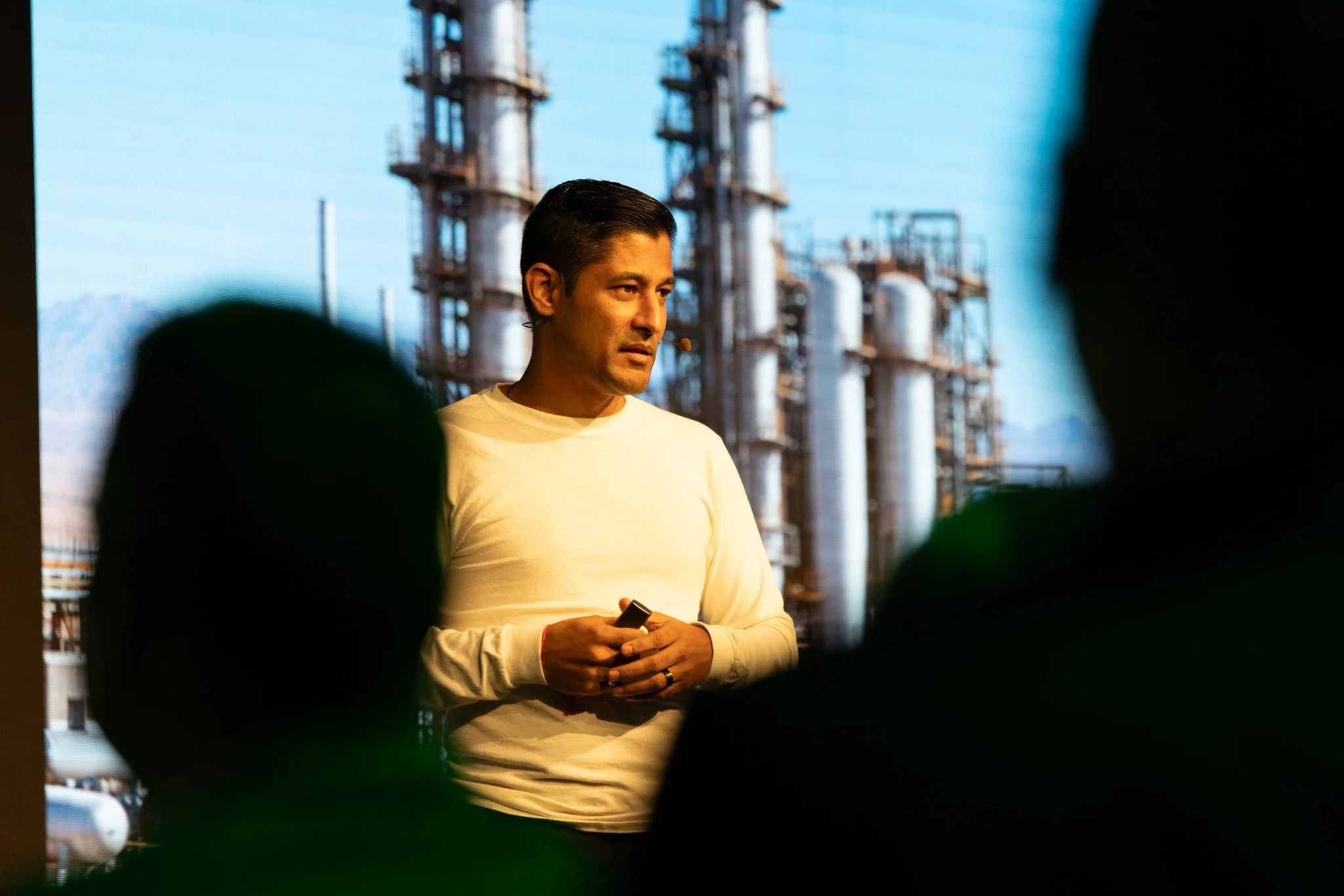 A man in a white long-sleeve shirt holds a microphone and stands in front of an industrial background with tall pipes and structures. The photo is taken through a dark frame, with some silhouettes in the foreground.