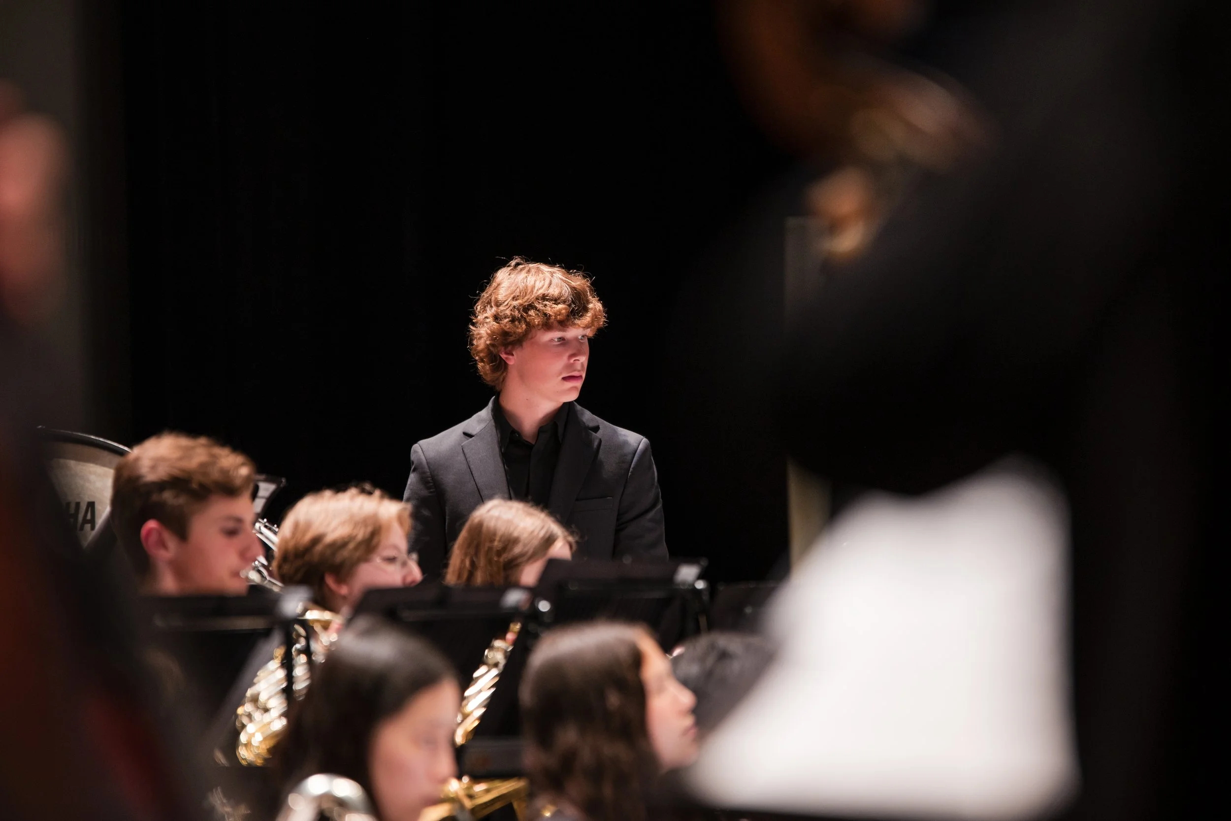 Young man with curly hair in a black suit attending a concert with a young orchestra, with several other musicians visible in the foreground.