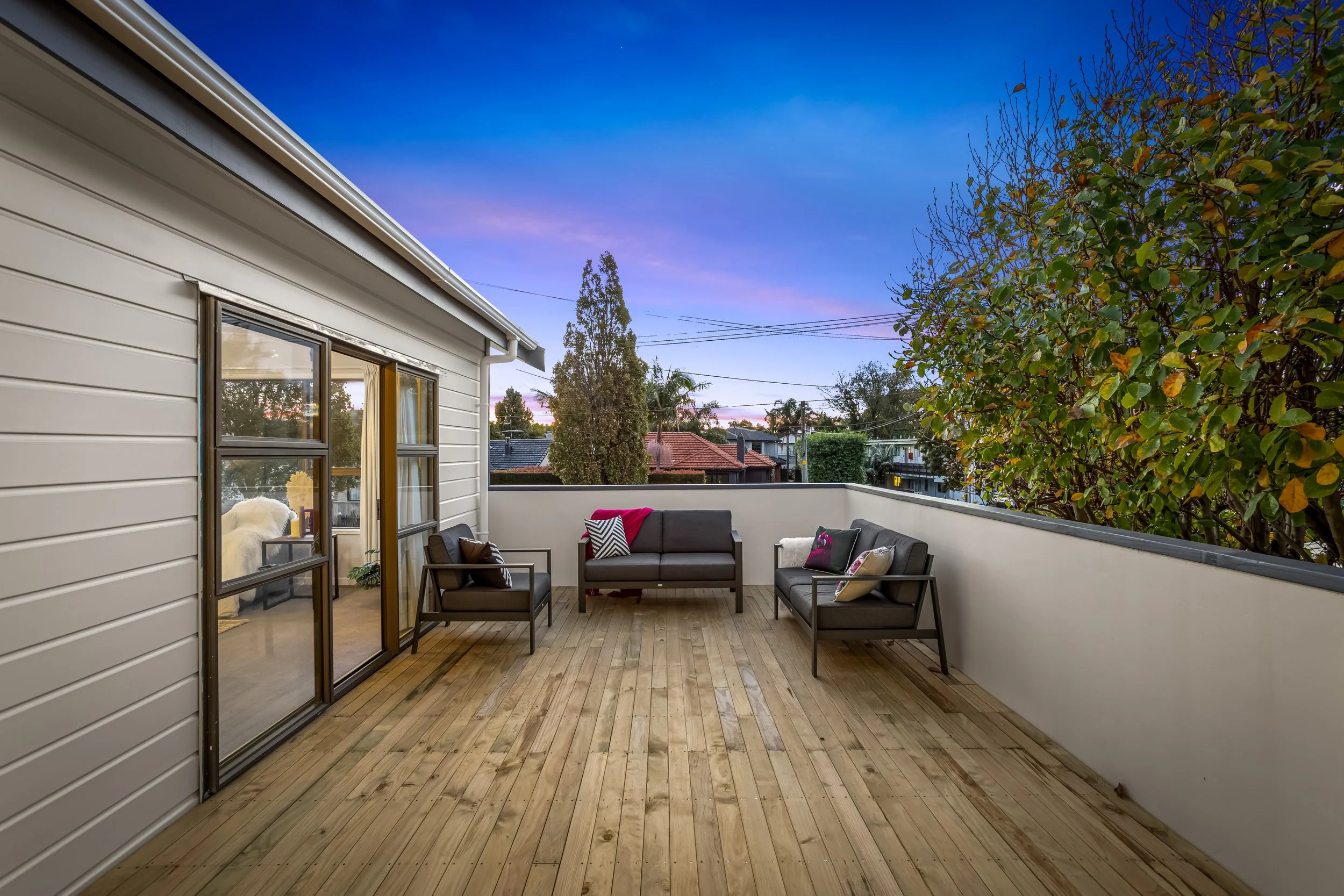 Outdoor balcony with wooden flooring, black outdoor furniture including a sofa and chairs with cushions, sliding glass door, white house siding, trees, houses, and power lines in the background during twilight.