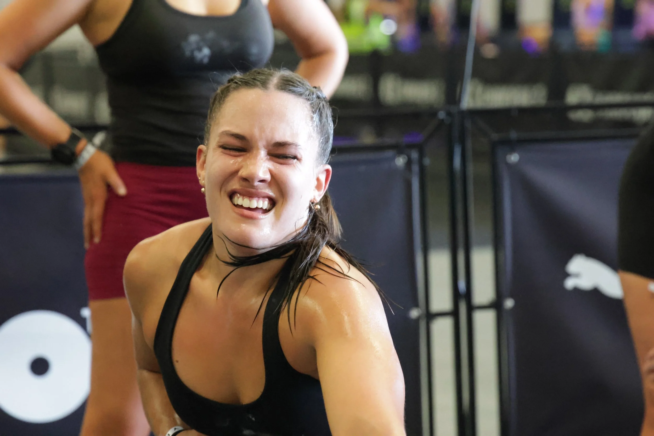A woman with wet hair and a black athletic top, smiling with her eyes closed, during a workout or fitness class.