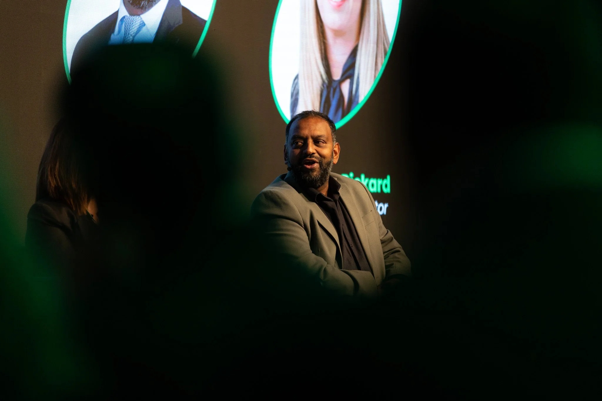 A man with a beard and dark hair wearing a gray blazer and black shirt, sitting and talking on a panel or at an event with a large screen behind him displaying images of two women.