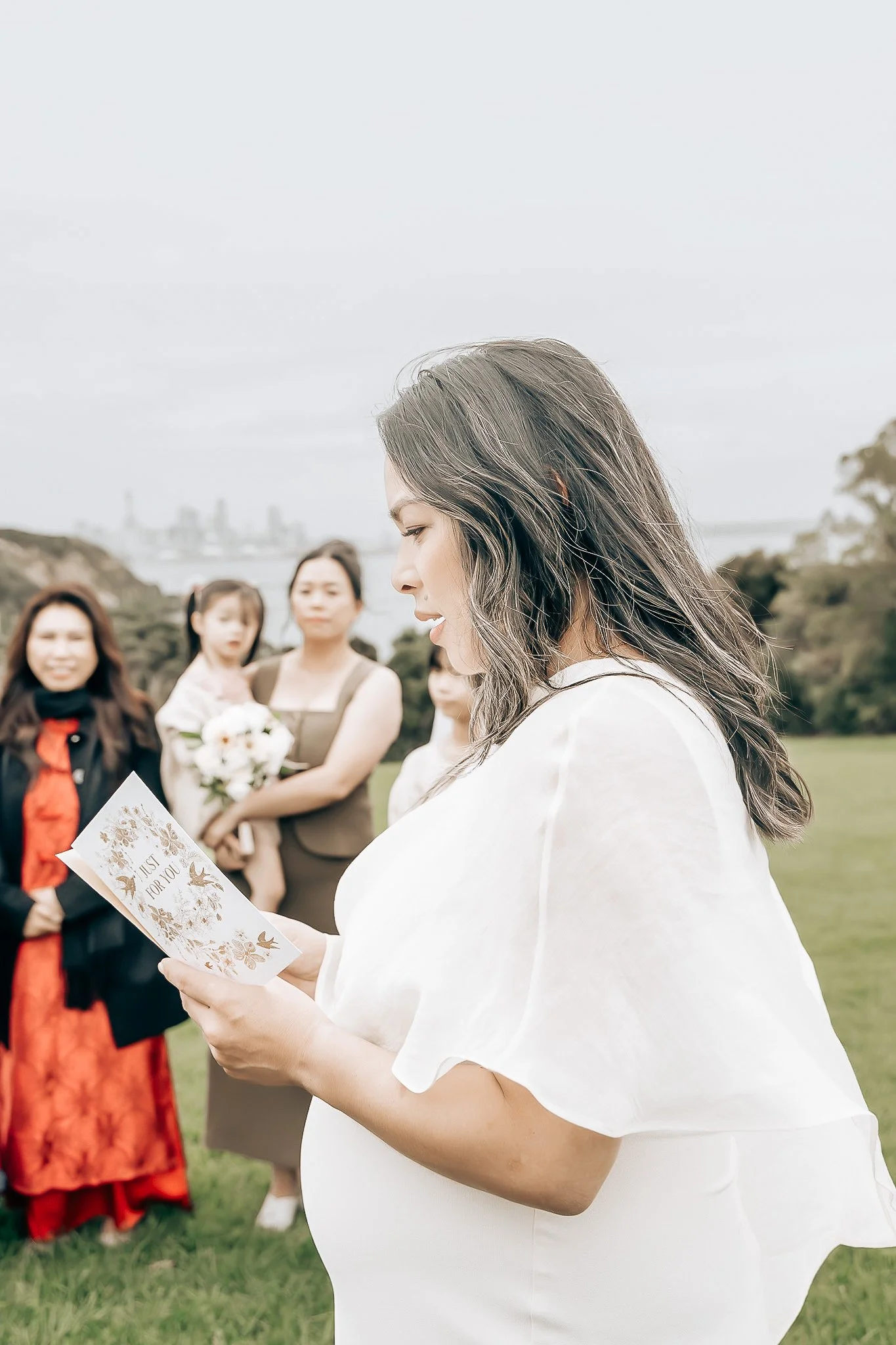 A pregnant woman with long wavy hair reading a card that says ‘Just for You’ at an outdoor gathering, with several women in the background on a grassy area with trees and a city skyline in the distance.