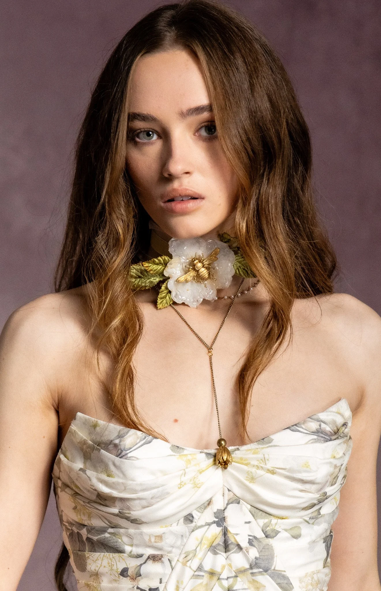 A woman with long brown hair wearing a floral strapless dress and a floral necklace with a bee and gold accents, standing against a muted purple background.