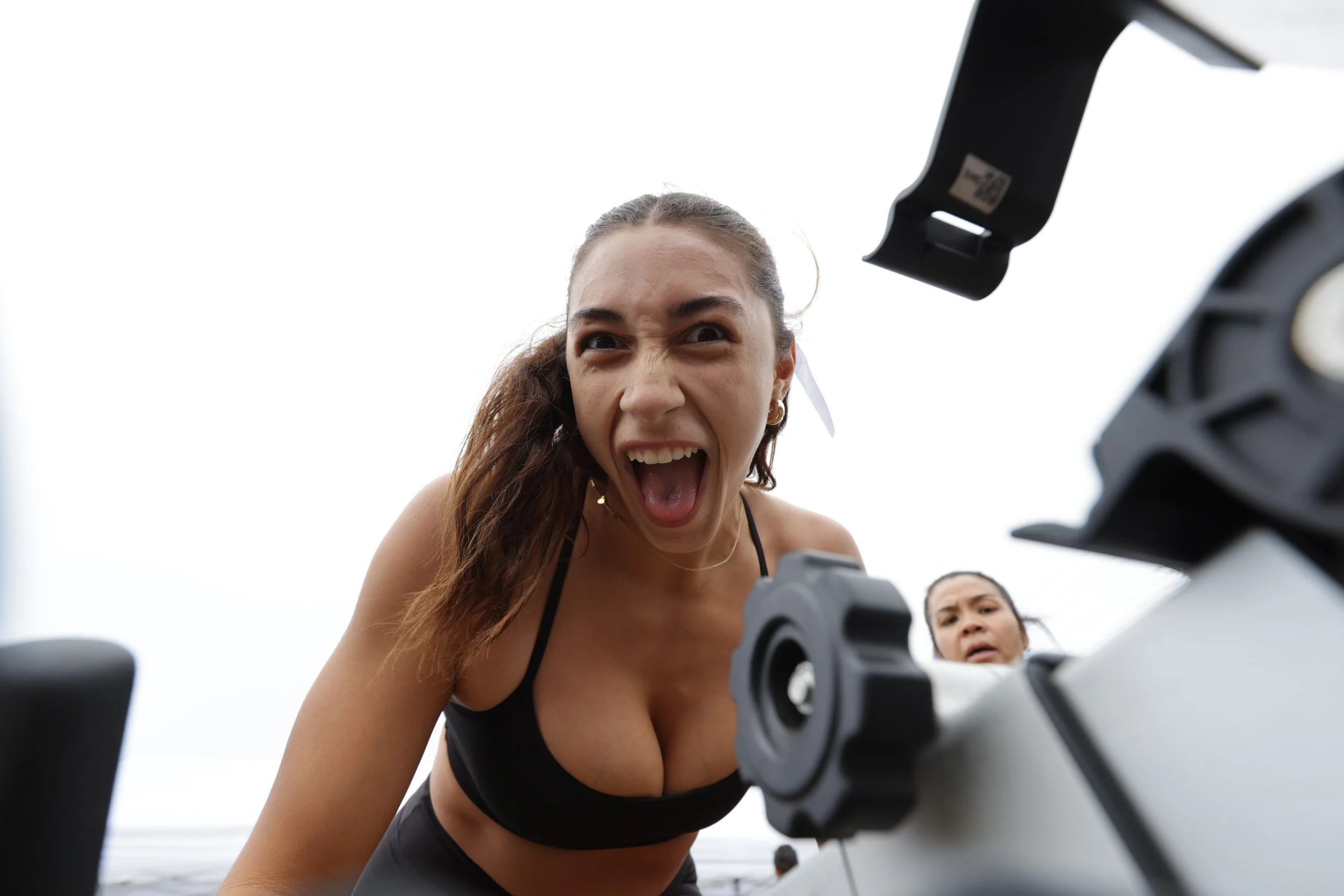 A woman with long hair and a black top is leaning over a drone, looking angry or excited, with her mouth open and eyes wide, while another woman stands behind her, looking concerned or surprised. The background appears to be outdoors with a cloudy sk