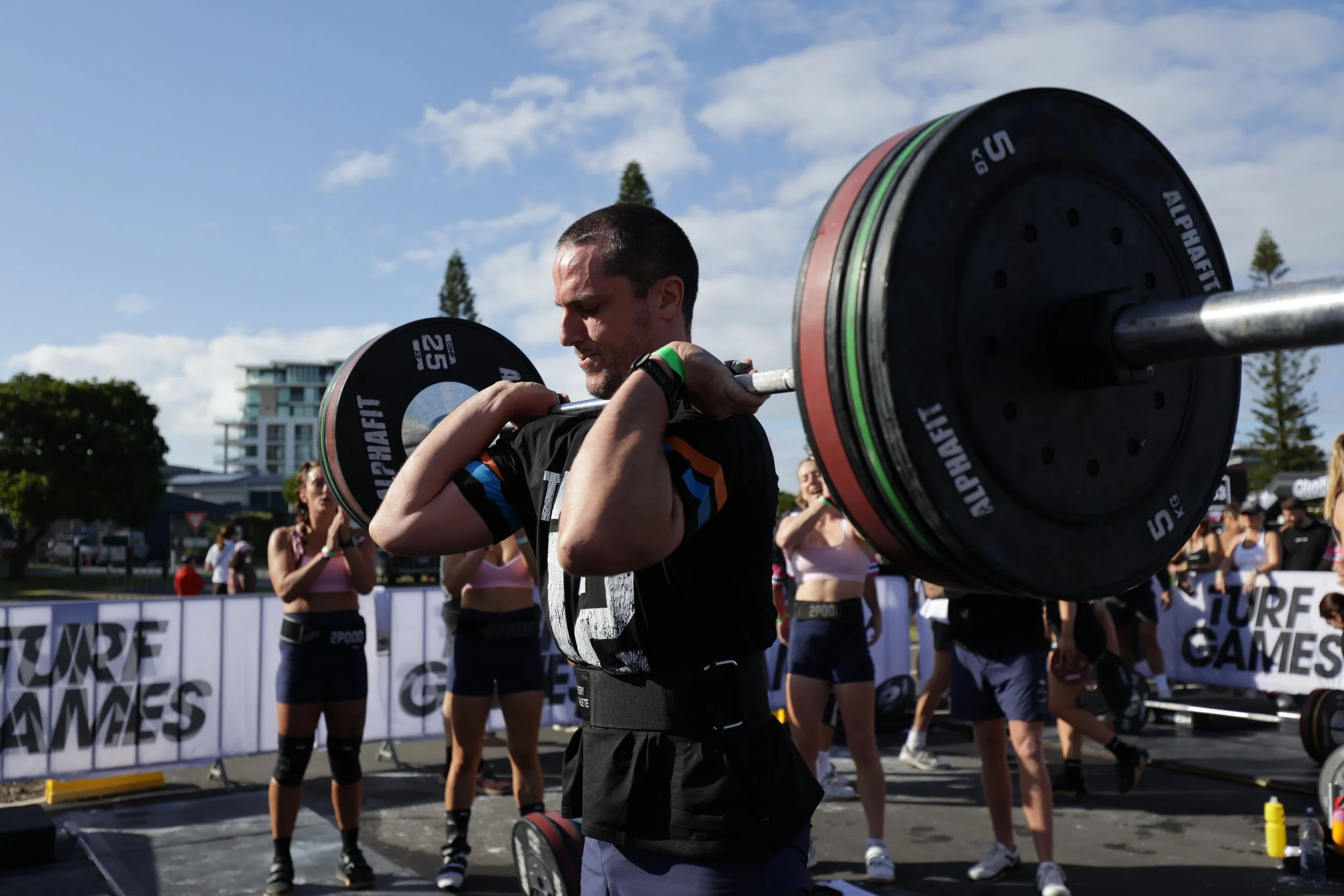 A man lifting a barbell during a fitness event outdoors with other women in workout attire in the background.