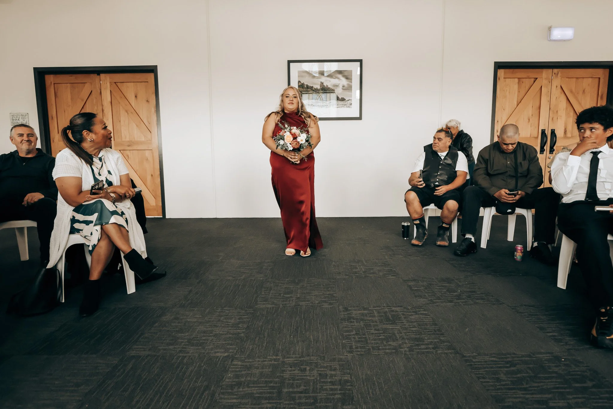 A woman in a red dress holding a bouquet of flowers stands in the center of a room with white walls and wooden doors, while five people sit on chairs around her, some looking at her and some using their phones.