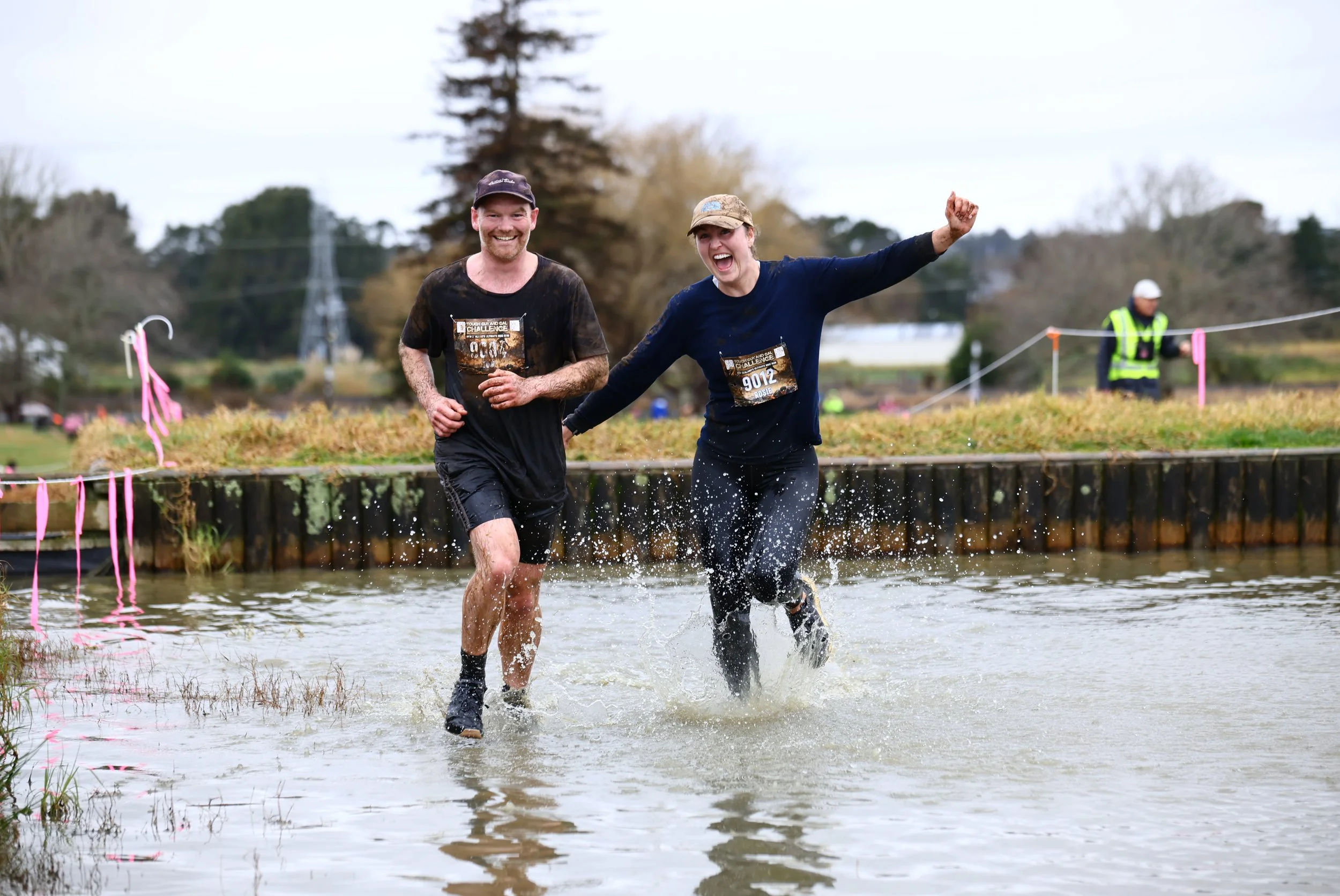 Two people running through a shallow water crossing during a muddy mud run race, smiling and splashing water on a cloudy day.