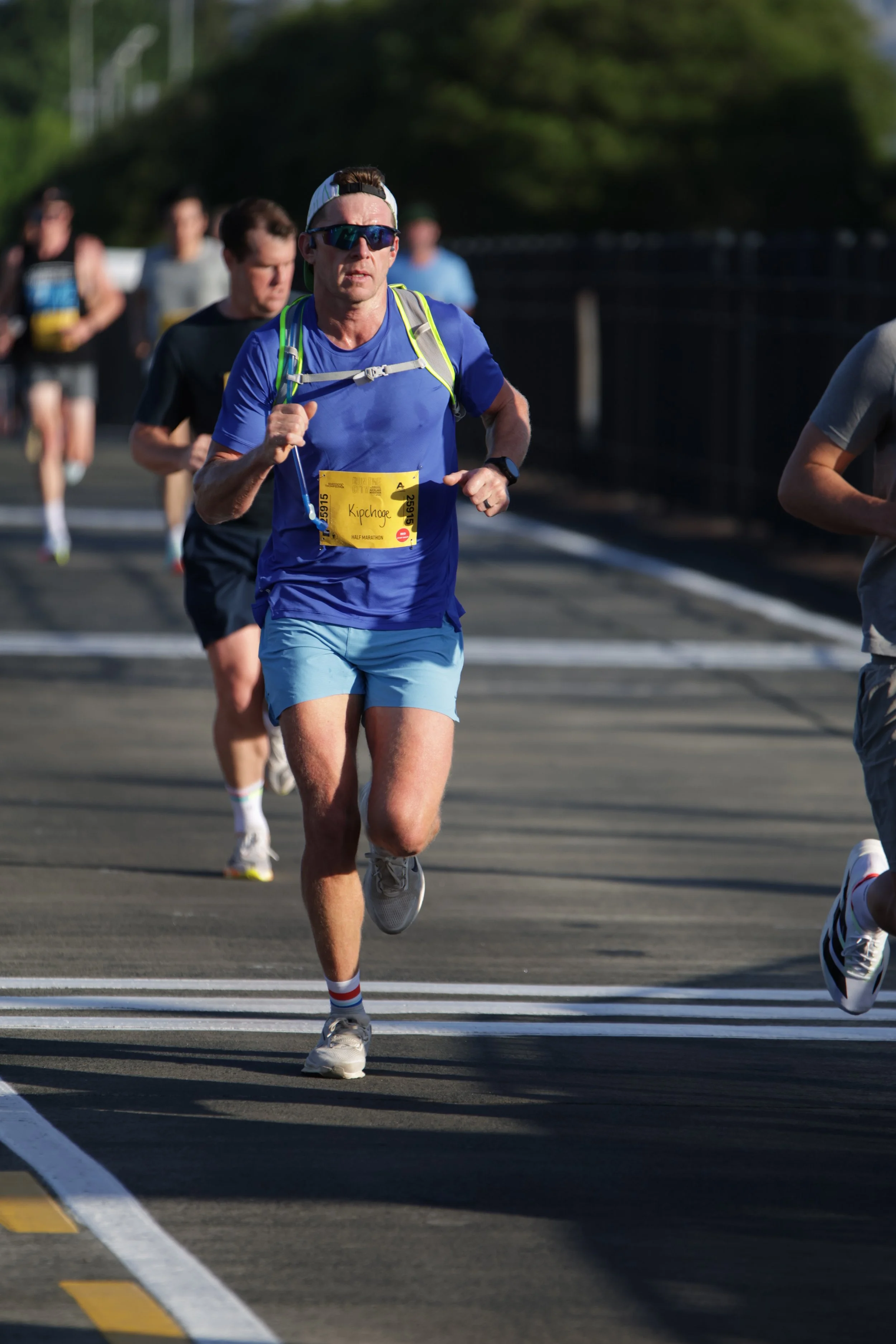 A group of runners participating in a marathon race on a sunny day, with the runner in the foreground wearing a blue shirt, light blue shorts, sunglasses, a white cap, and a yellow race bib.
