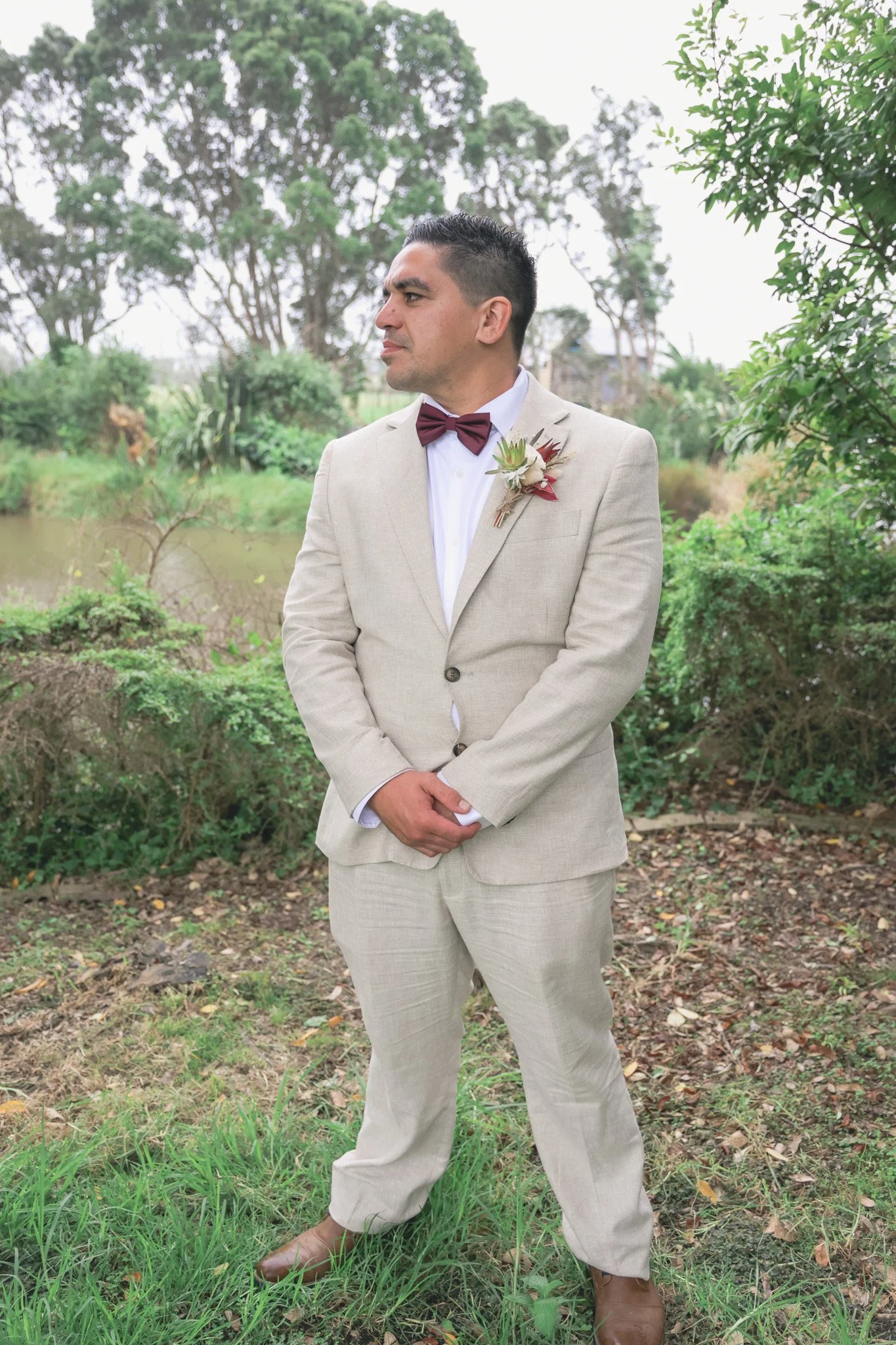 A man dressed in a beige suit with a burgundy bow tie and boutonniere, standing outdoors near a body of water with trees in the background.