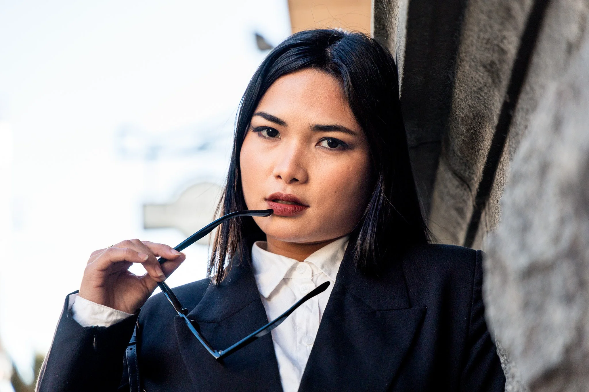 A young woman with dark hair and makeup wearing a black blazer and white shirt, holding sunglasses, standing against a textured stone wall.