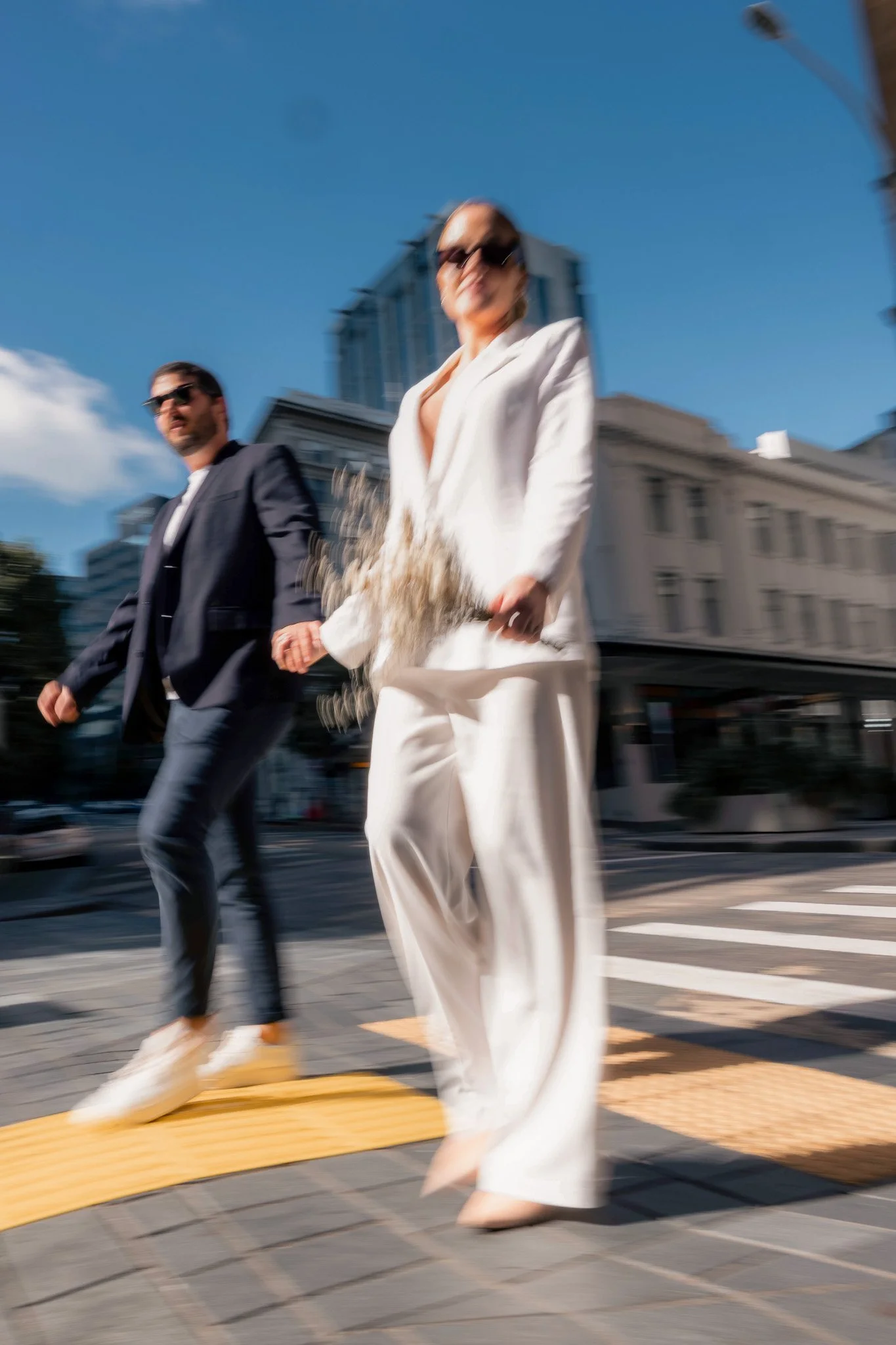 Two stylish individuals walking on a city street, with a woman in a white suit and sunglasses, and a man in a dark suit, both holding hands, with tall buildings, blue sky, and motion blur indicating movement.