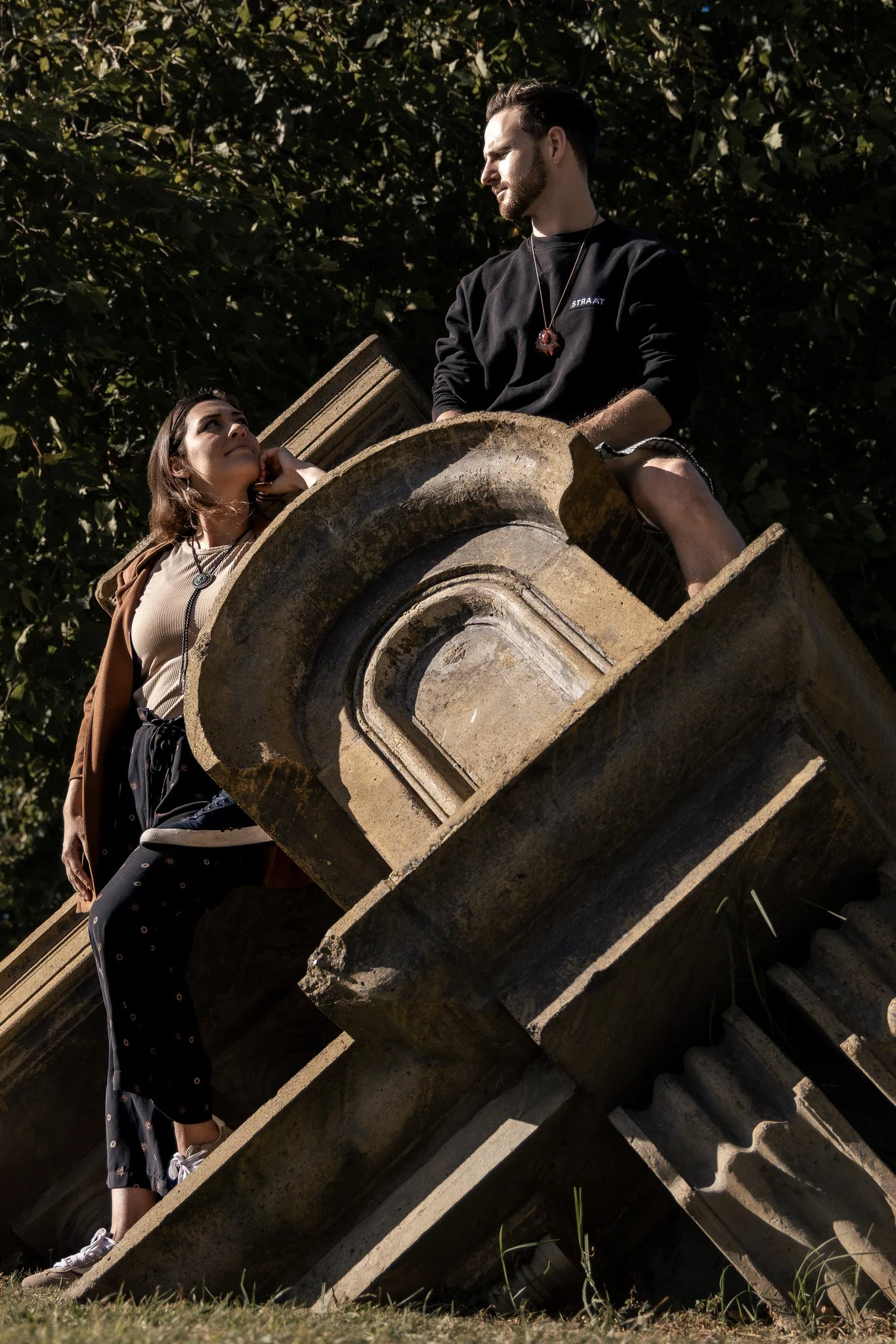A man and a woman sitting on a large, old stone architectural detail outdoors surrounded by lush greenery, with sunlight casting shadows.