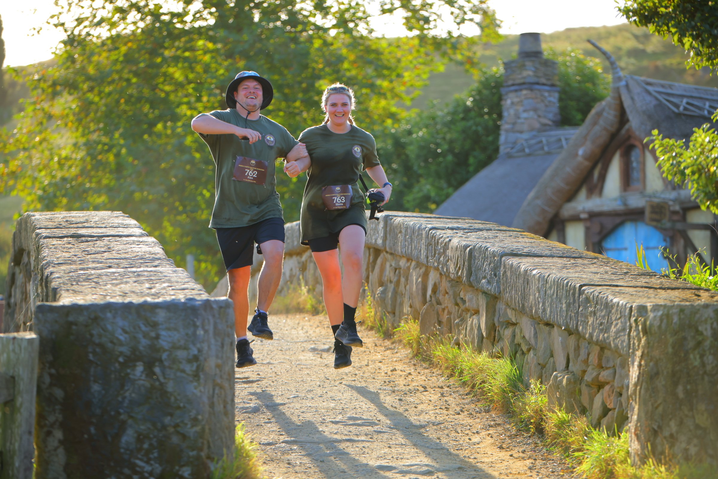 Two people, a man and a woman, running or jogging on a stone bridge in a park or natural setting with trees and sunlight. They are wearing dark green shirts and black shorts, and are smiling.