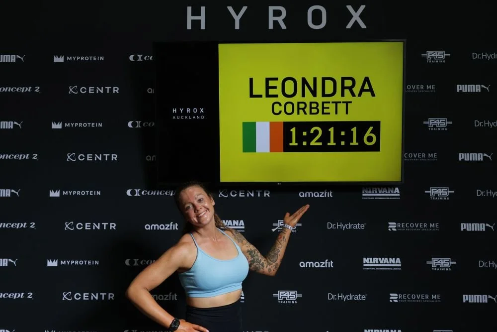 A smiling woman in a blue sports bra standing in front of a black banner with various logos, pointing to a digital screen displaying a name, time, and flags.