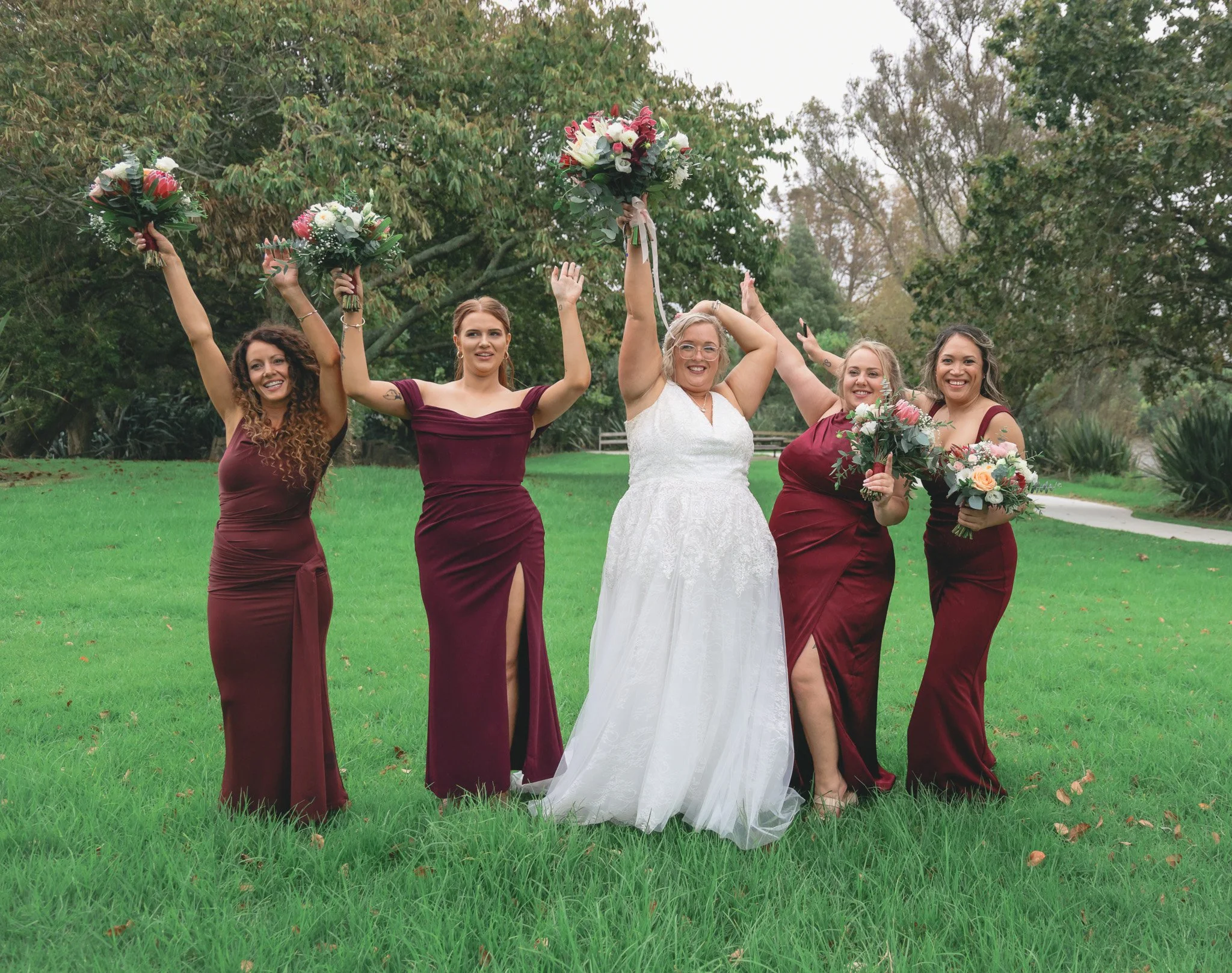 A group of five women celebrating outdoors on a grassy area with trees in the background, holding bouquets of flowers, dressed in formal attire, with the woman in the center wearing a white wedding dress.